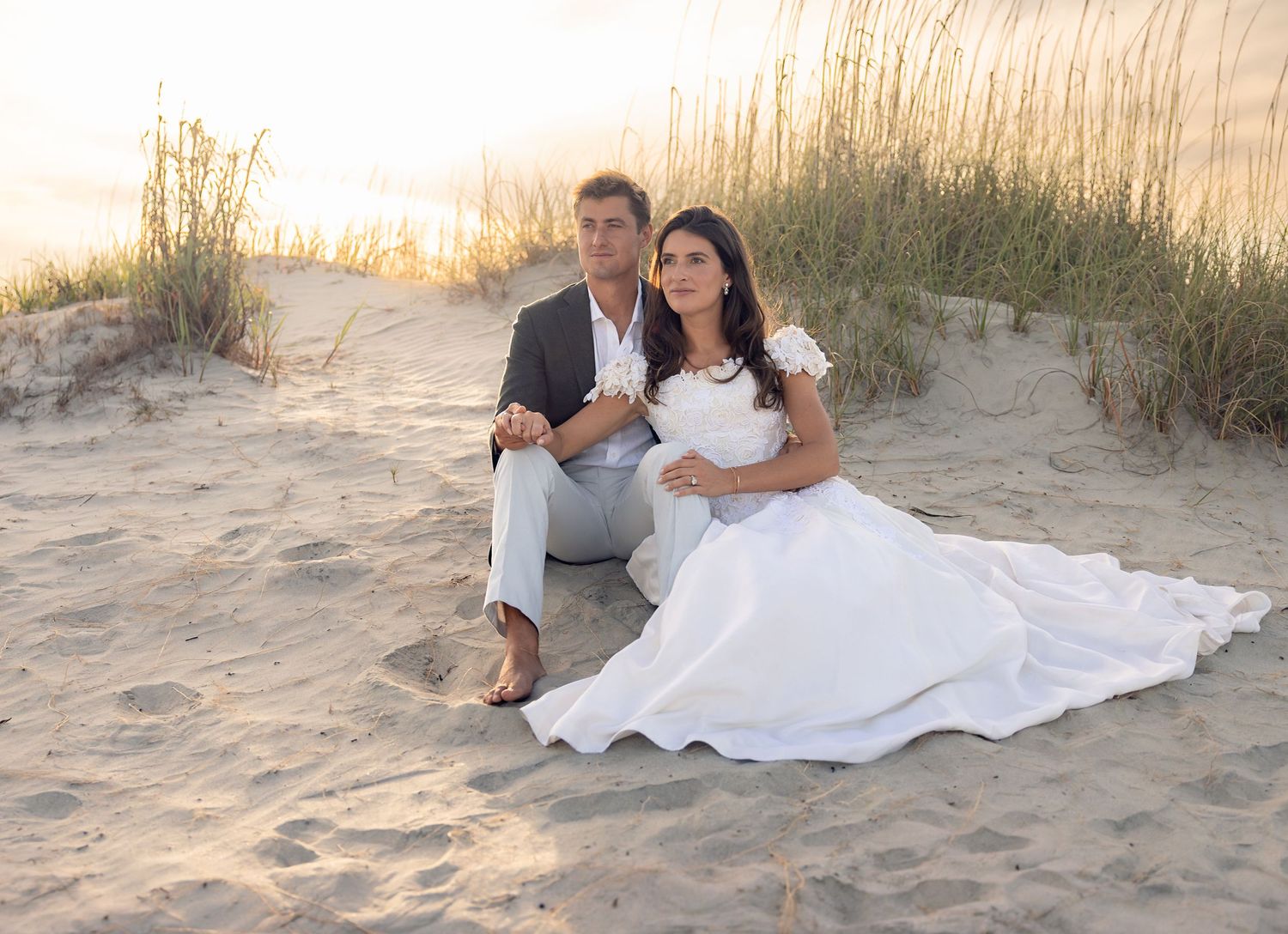 Couple in wedding attire sitting on sandy beach dunes at sunset, bride in white gown beside groom in gray suit.