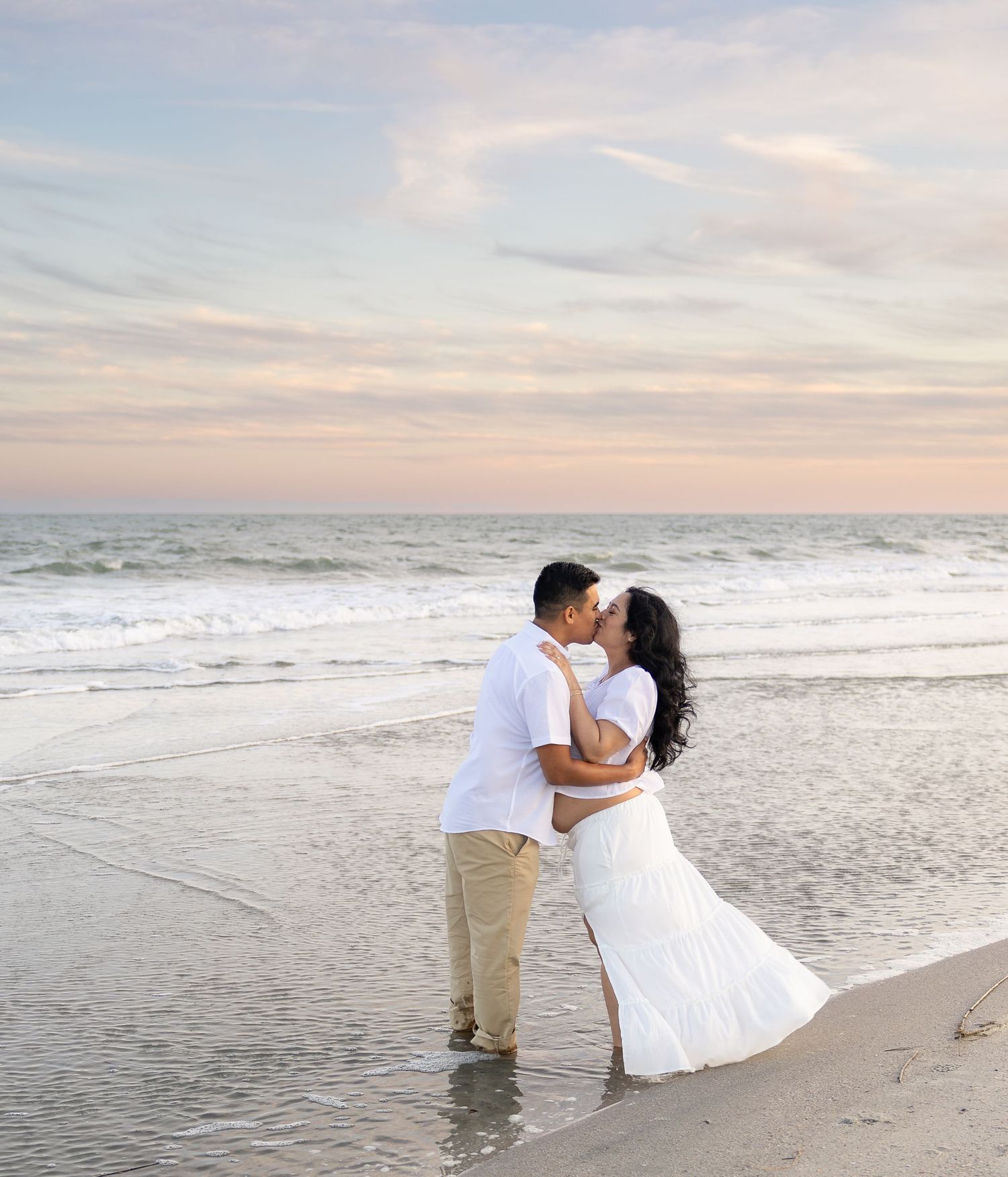 Couple kissing on a beach at sunset, wearing white outfits, with ocean waves and pastel sky in the background.