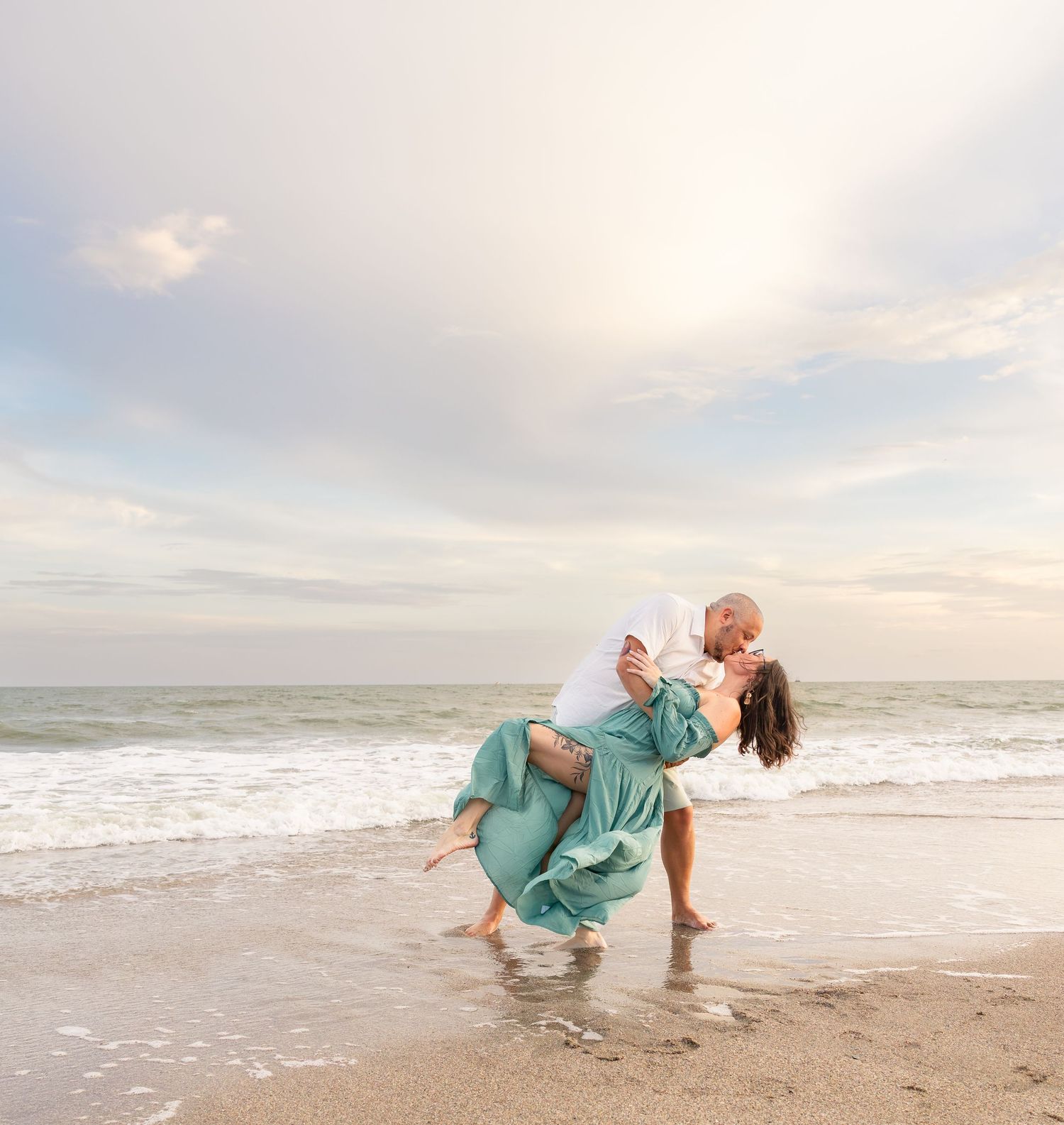 Couple sharing a romantic dip kiss on a sandy beach at sunset, woman in flowing teal dress, waves in background.