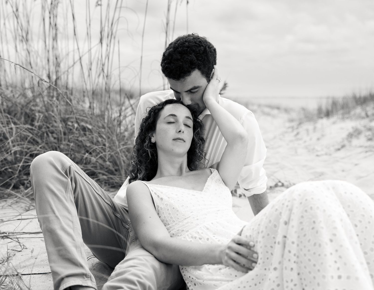 Black and white photo of a romantic couple sitting on a sandy beach path, woman leaning back on man.
