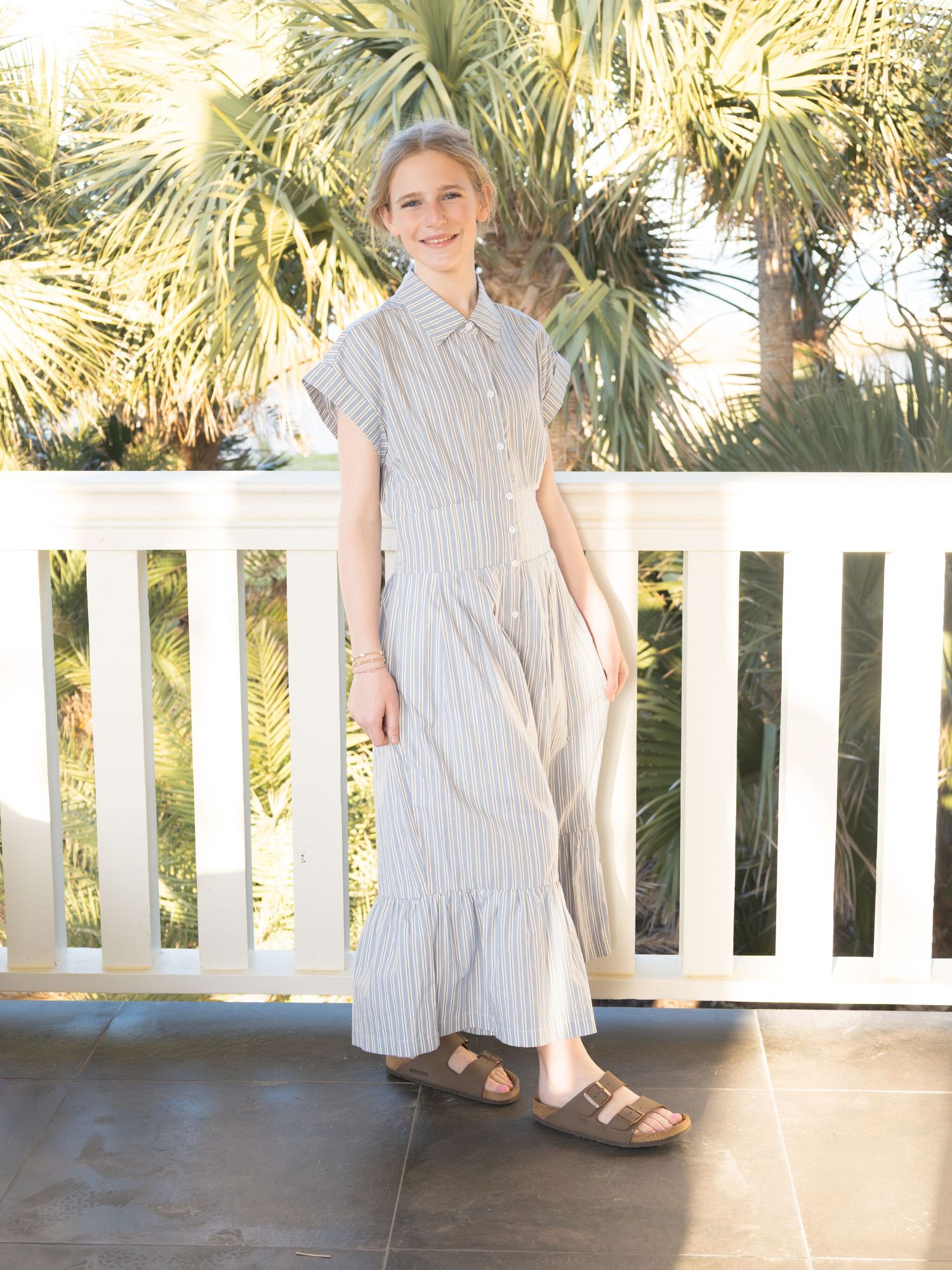 A woman wearing a light-colored dress stands on a porch with palm trees in the background.