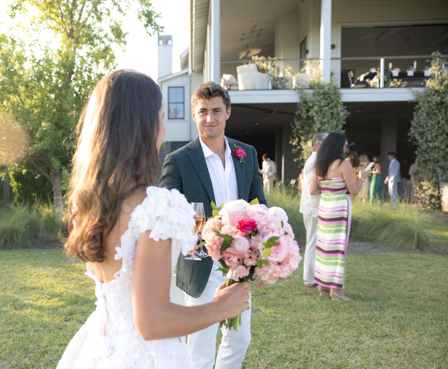 Bride holding pink bouquet faces groom in dark suit at outdoor wedding reception on green lawn.