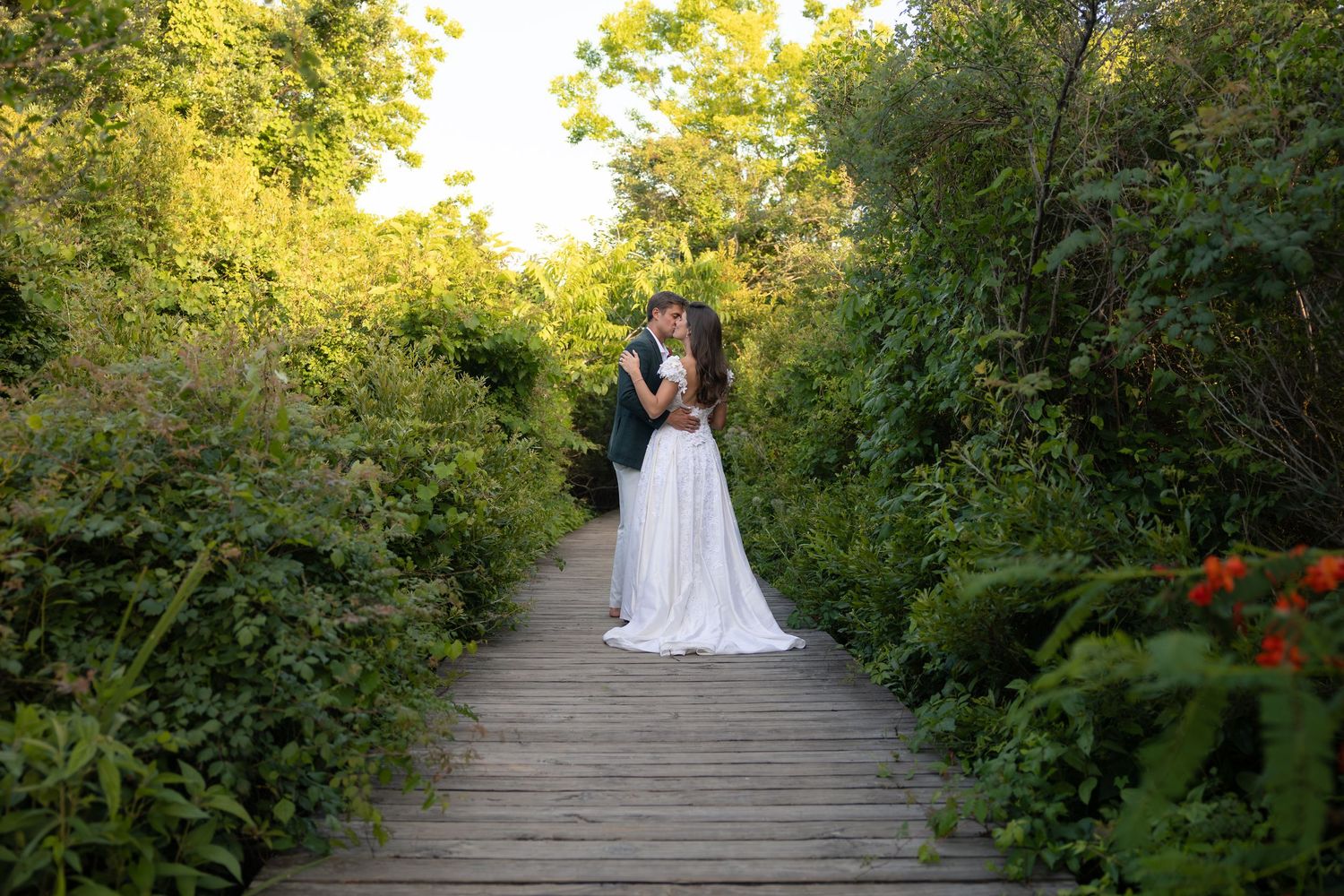 Bride and groom embracing on a wooden boardwalk path surrounded by lush green garden foliage on their wedding day.