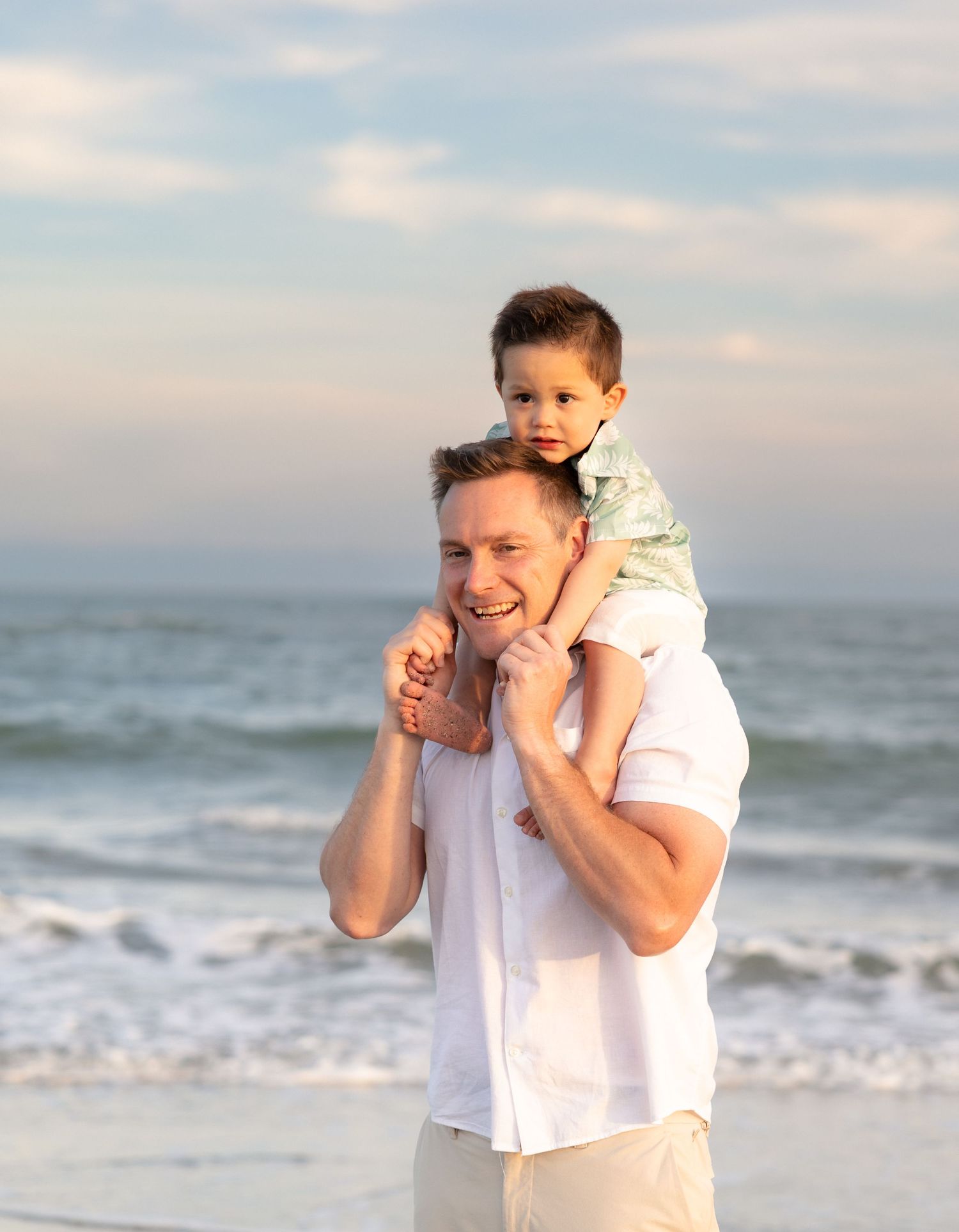 Smiling father carrying young son on shoulders at the beach with ocean waves behind them.