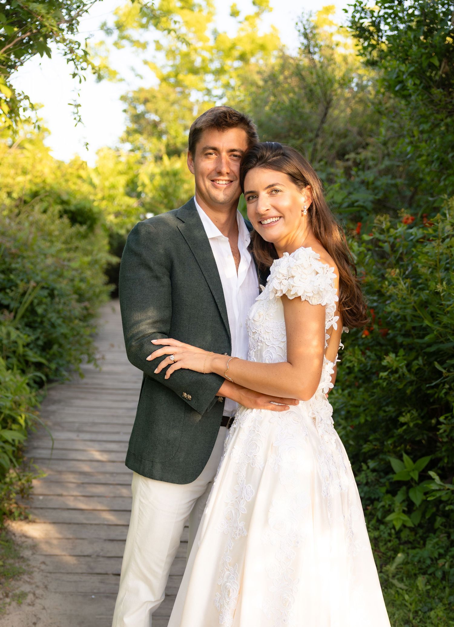 A couple in wedding attire embraces on a wooden path surrounded by lush greenery in a romantic outdoor setting.