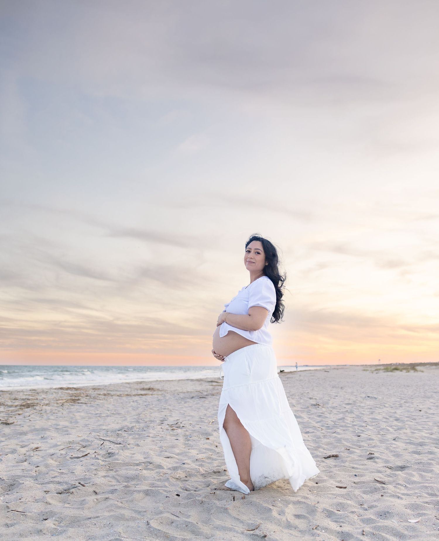 Pregnant woman in white dress posing on sandy beach at sunset, cradling baby bump with ocean waves in background.