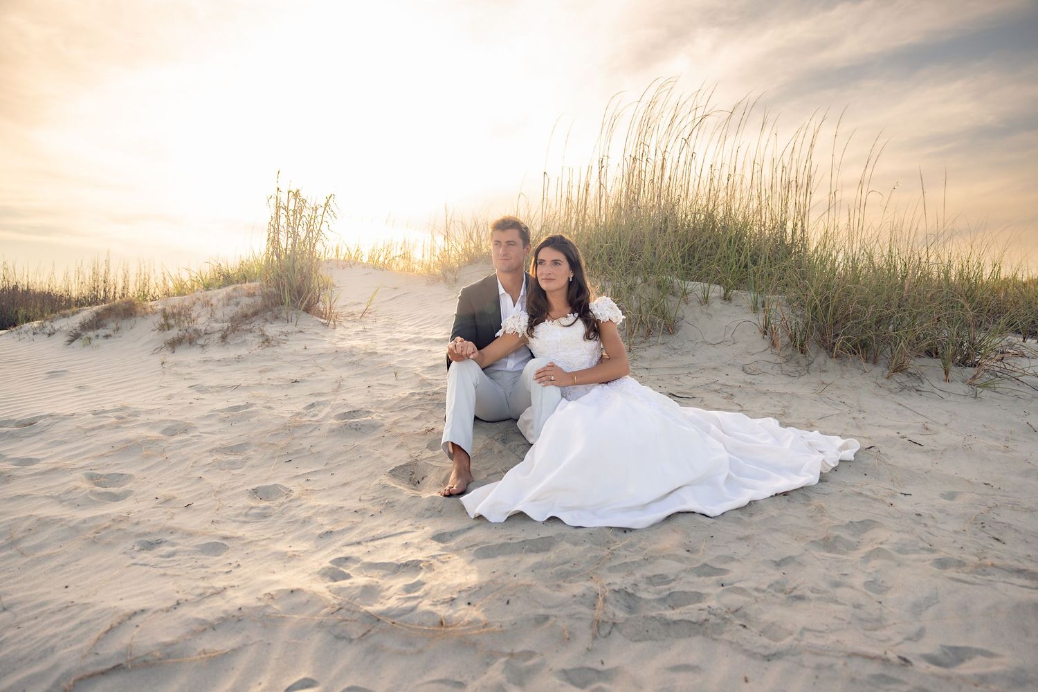Couple in wedding attire sitting on sandy beach dunes at sunset with sea oats in background.