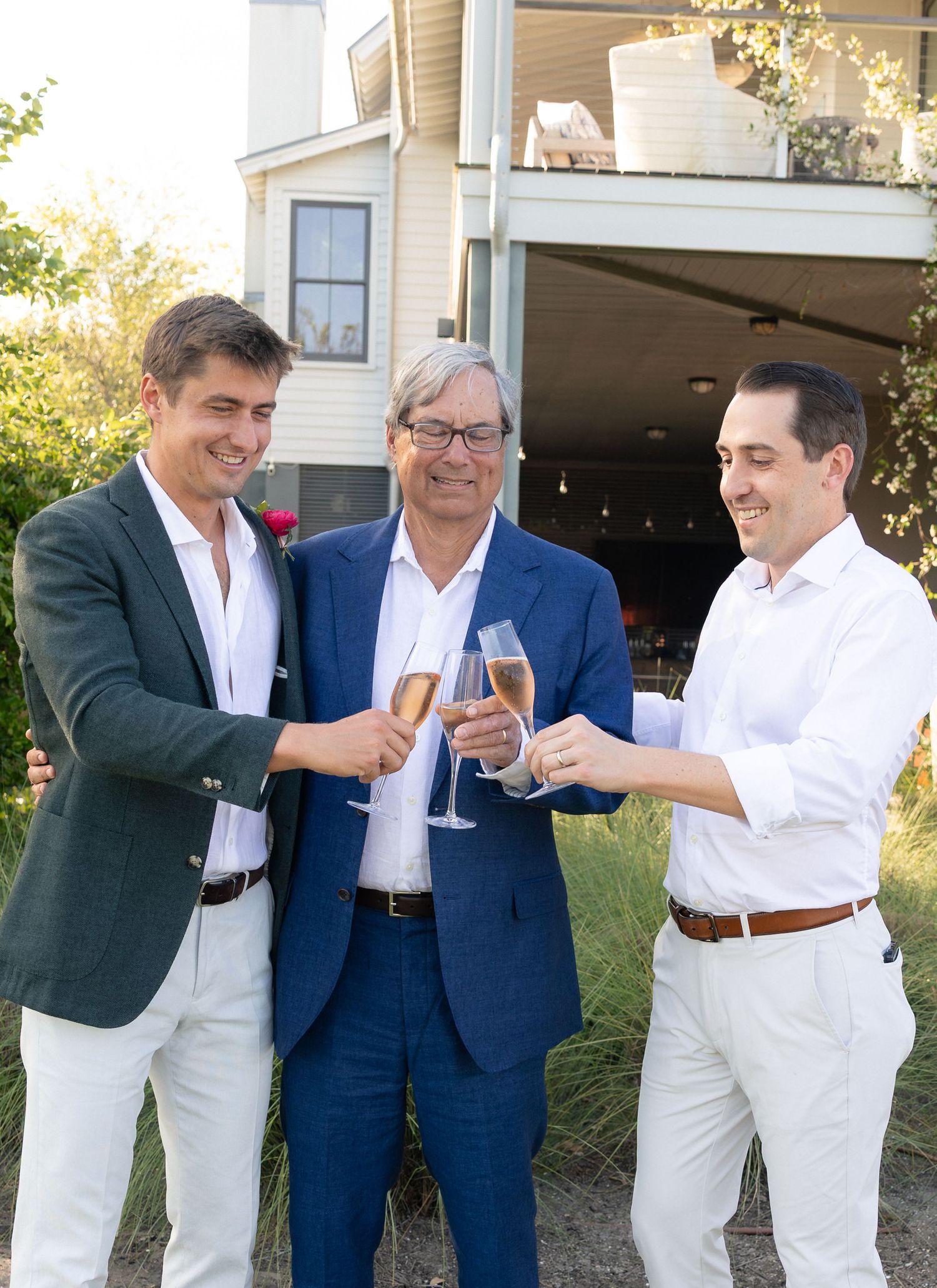 Three men in elegant attire toasting with champagne glasses outdoors near a house.