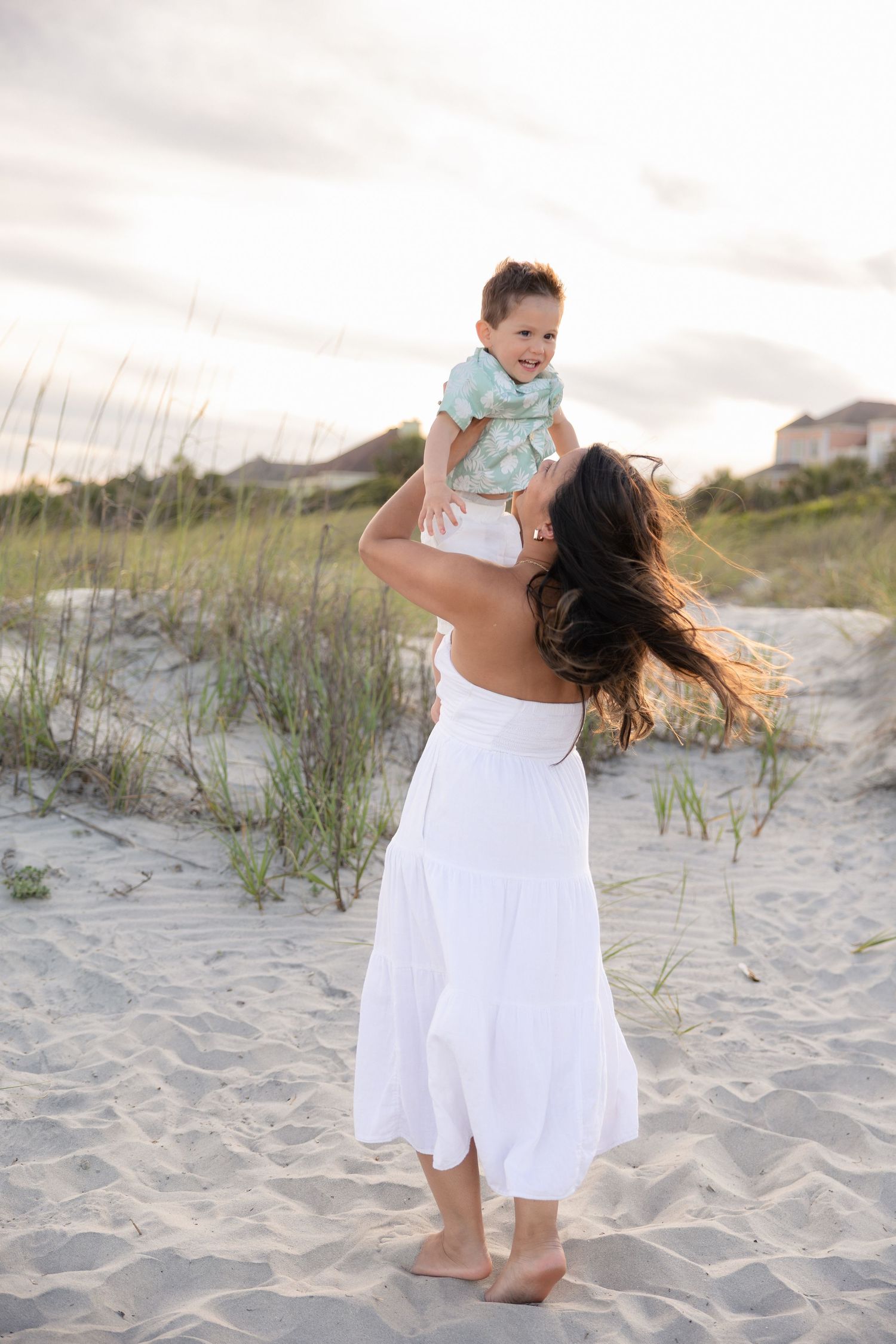 A mother in a white dress lifts her smiling baby on a sandy beach with dunes and grass in the background.