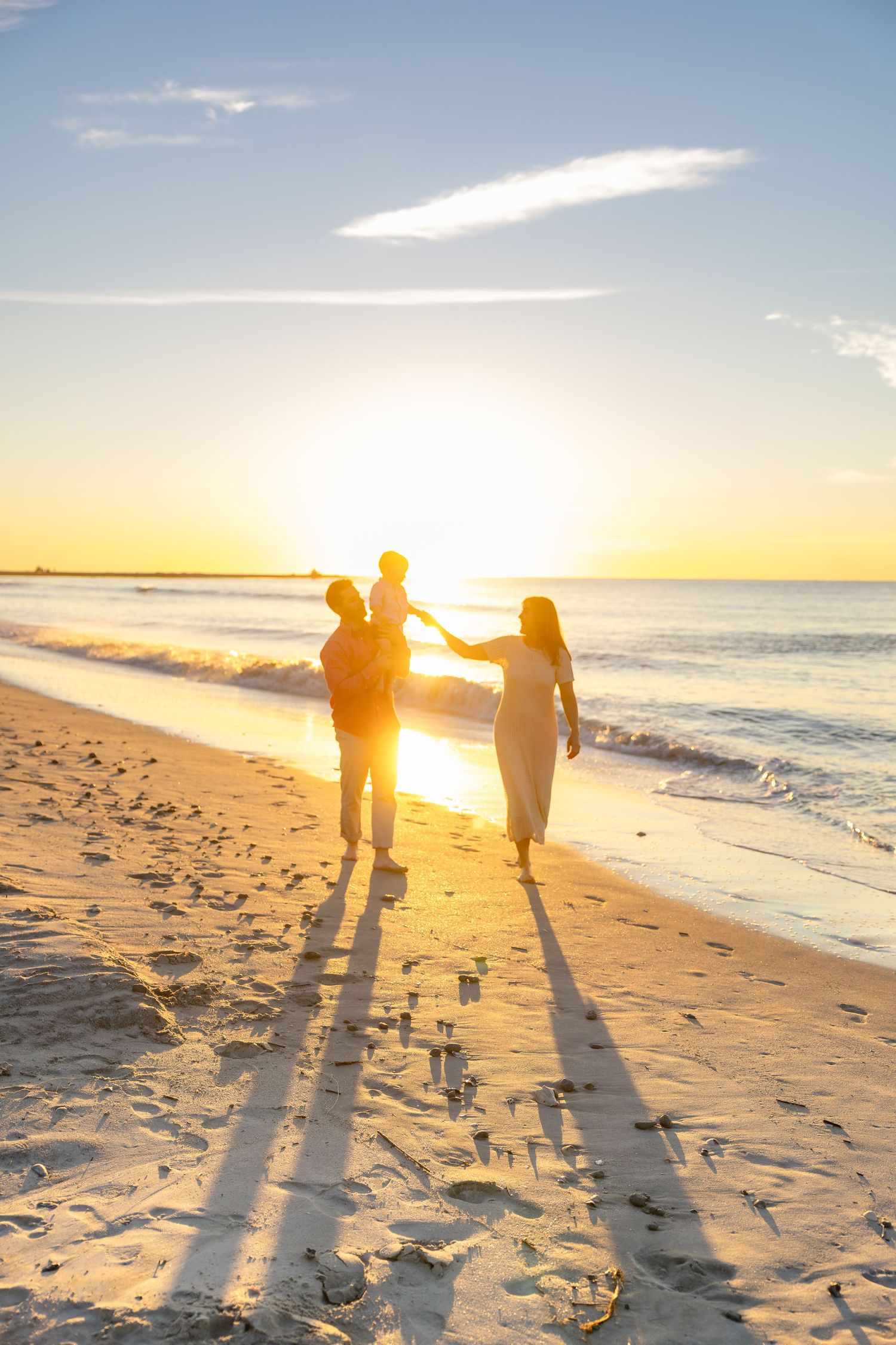Family of three walking hand in hand along a sandy beach at golden sunset, casting long shadows beside gentle ocean waves.