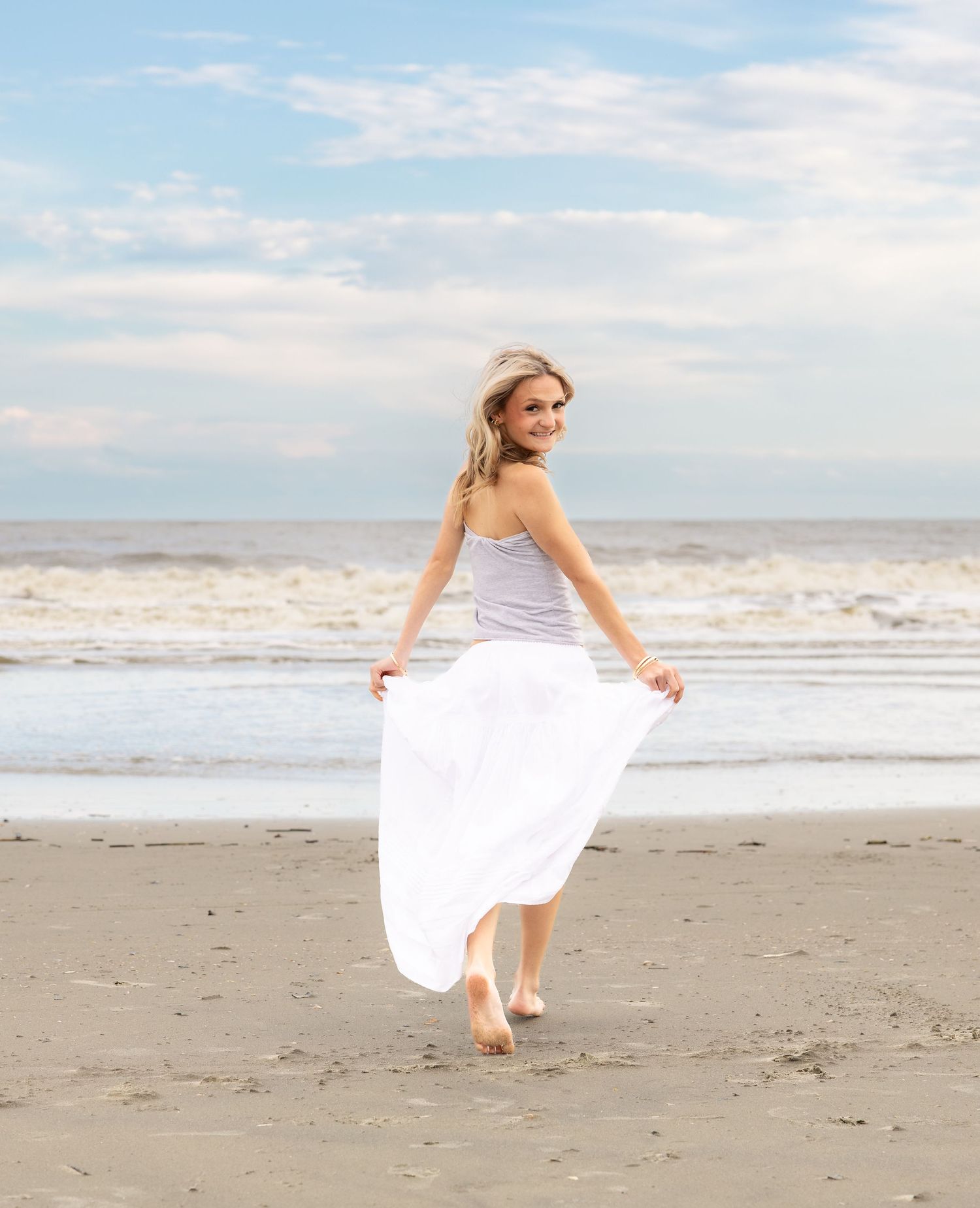 Young blonde woman in white skirt and gray top walking barefoot on sandy beach with ocean waves behind her.
