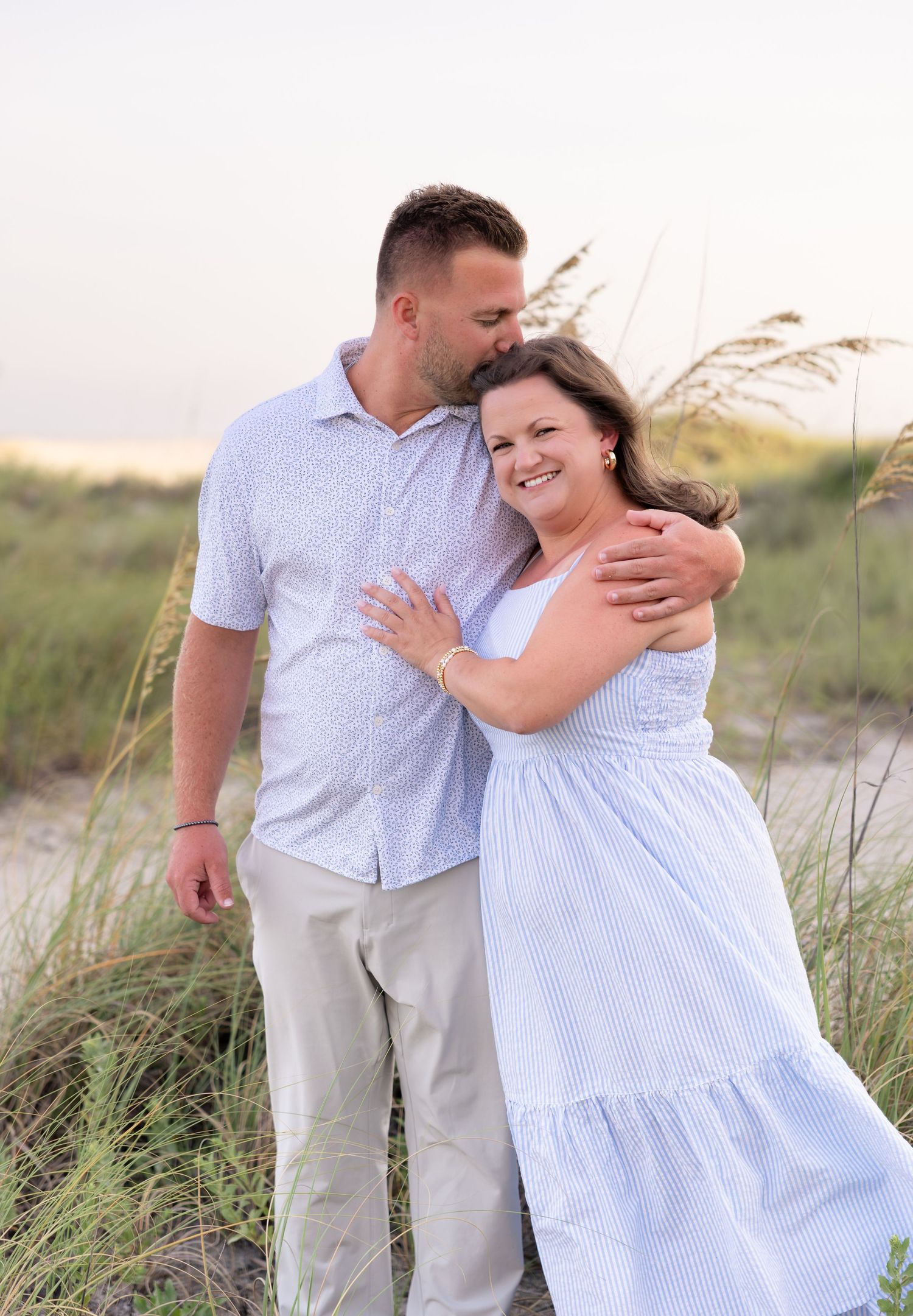 Couple embracing on a sandy beach at sunset, woman in blue dress smiling, man in white shirt kissing her head.