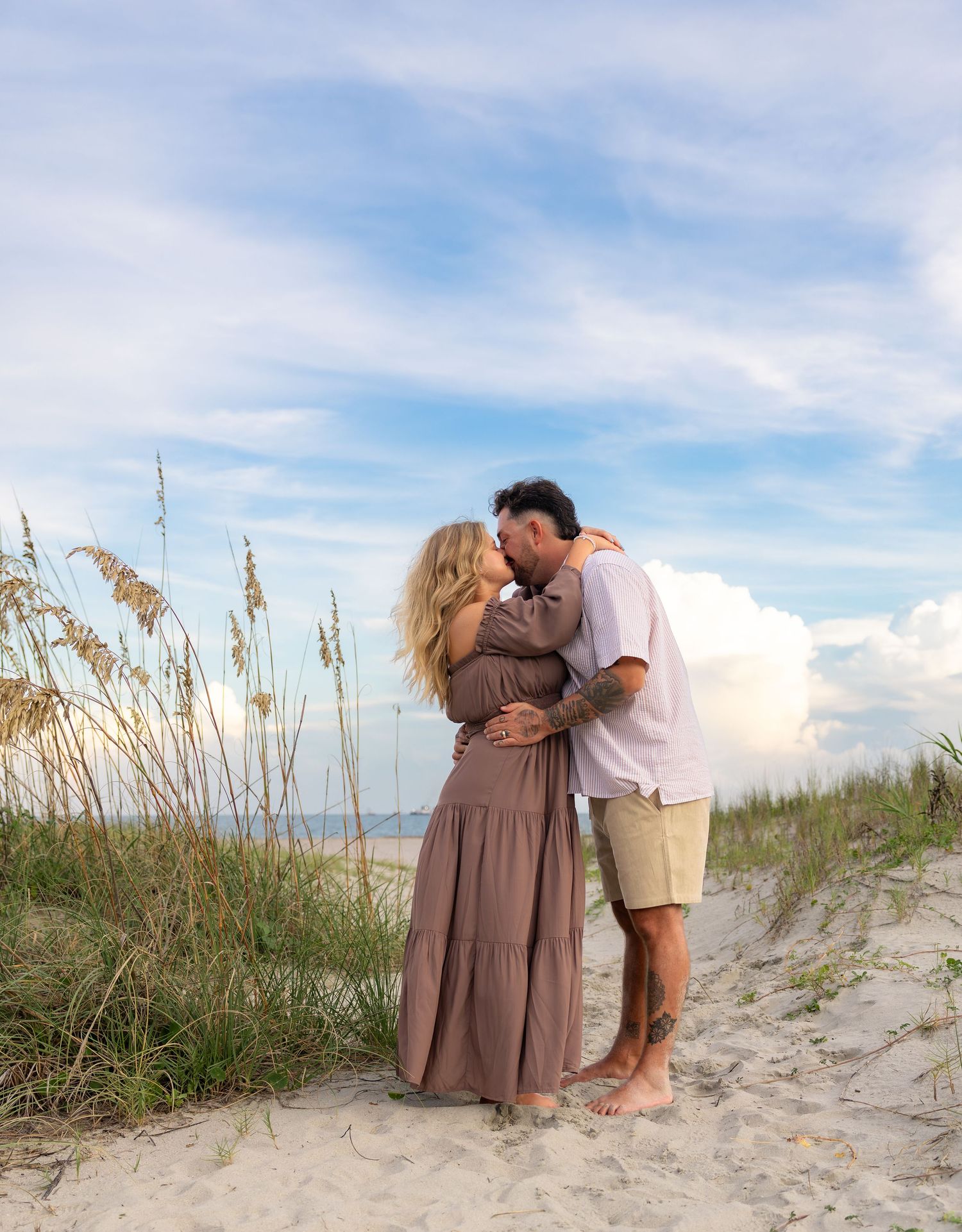 Couple embracing and kissing on a sandy beach dune with sea oats and a soft blue sky with clouds in the background.