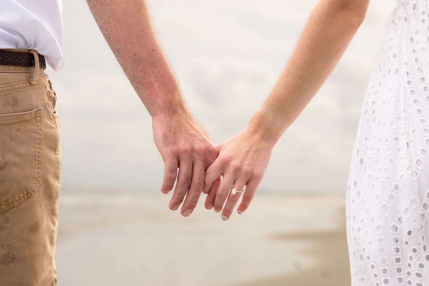 Couple holding hands on beach, woman wearing gold ring, romantic seaside engagement photo.