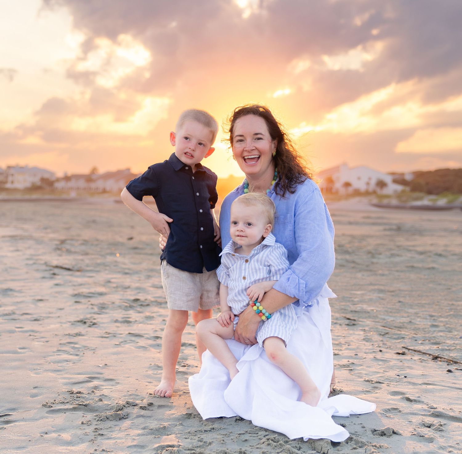 A woman and two young boys smile on a sandy beach at sunset with a colorful sky backdrop.