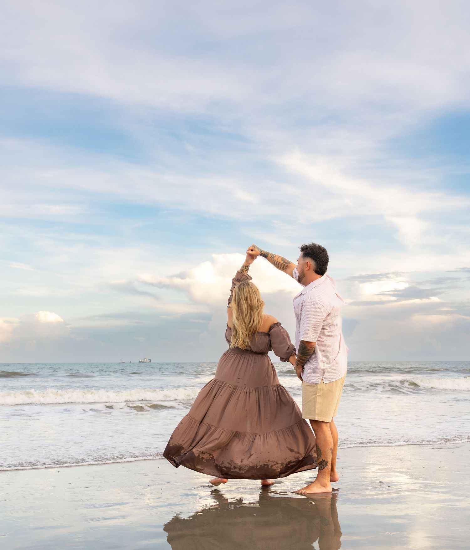 Couple dancing on a beach at sunset, woman in flowing brown dress being twirled by man in white shirt.