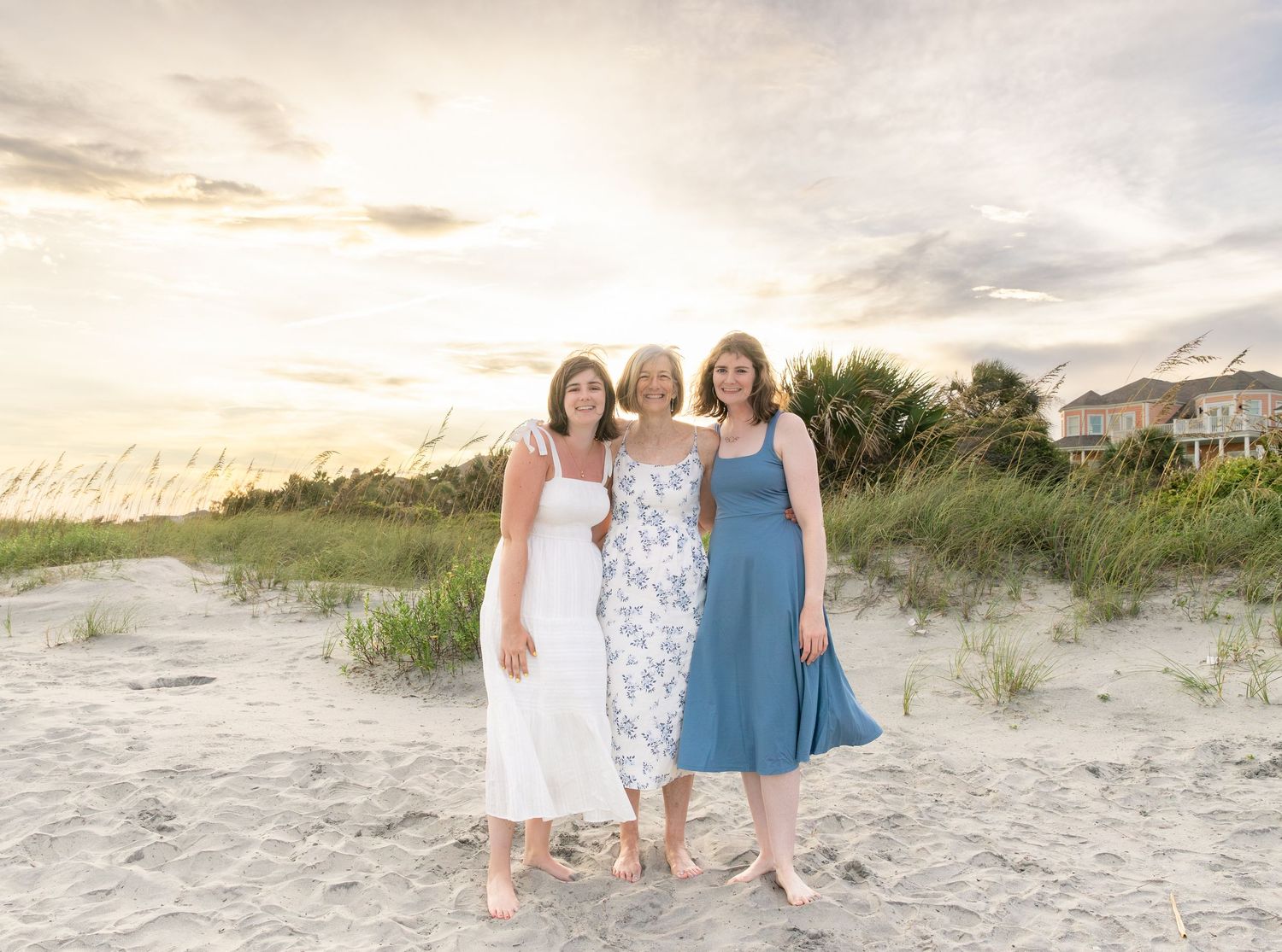 Three women in summer dresses pose together on a sandy beach at sunset with sea oats and coastal homes in background.