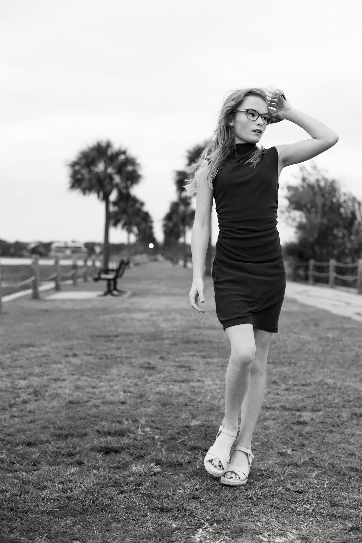 Black and white portrait of a young woman in glasses and a sleeveless dress posing on a grassy park path with palm trees behind her.