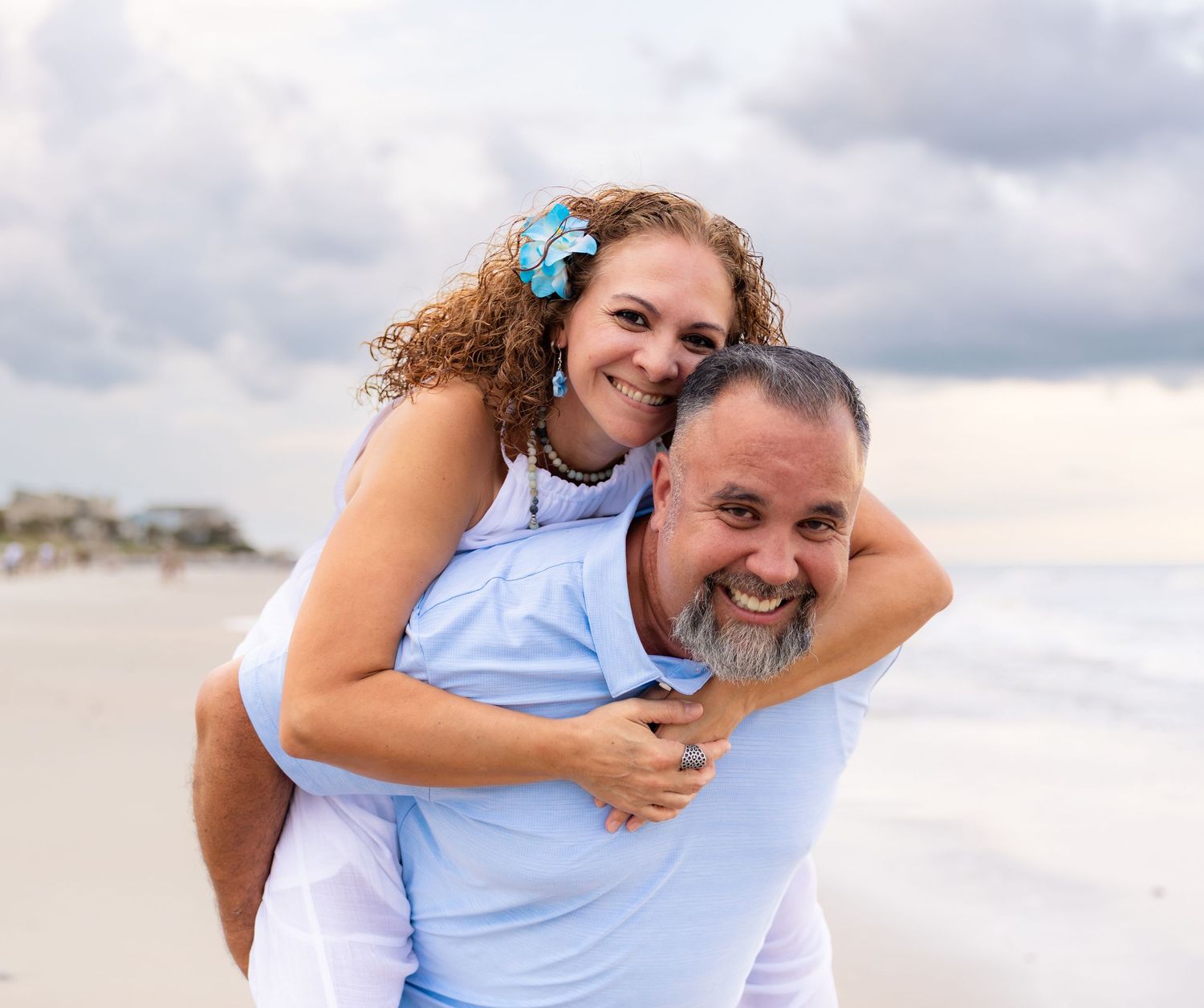 A happy couple poses on a sandy beach, woman on mans back, both smiling, cloudy sky backdrop.