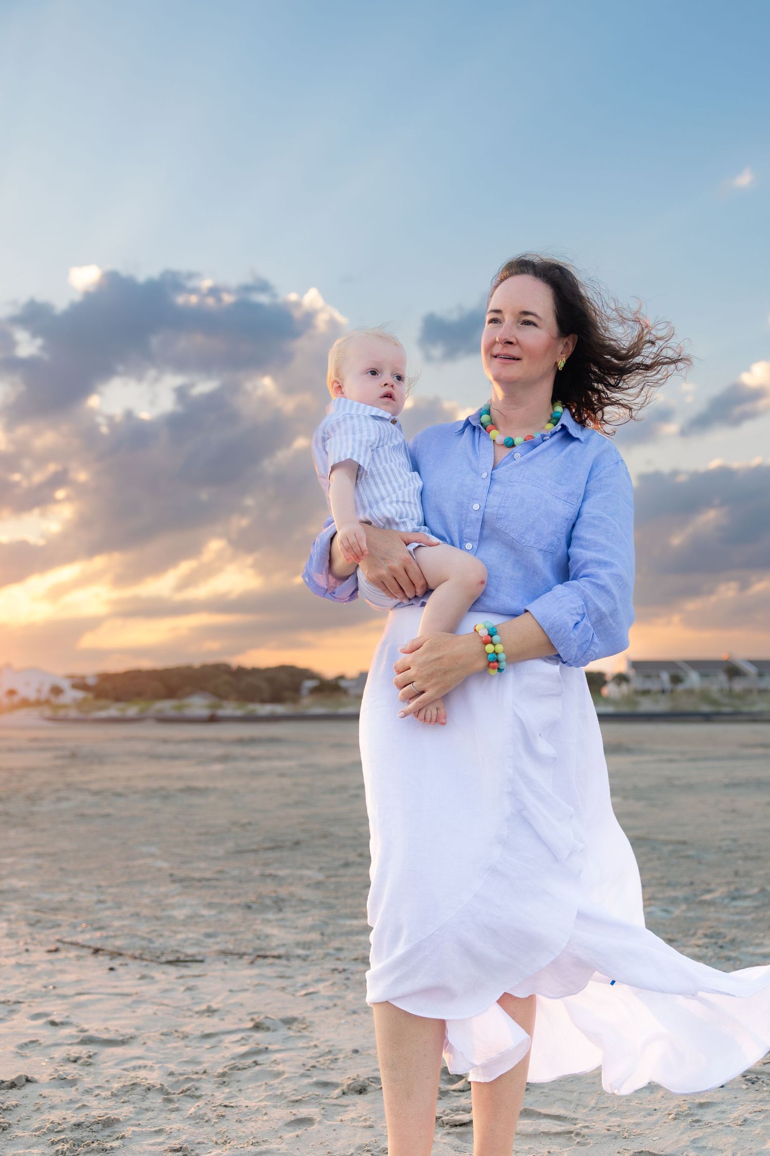 Mother holding baby on beach at sunset, wearing blue shirt and white skirt, with colorful necklace and wind-blown hair.