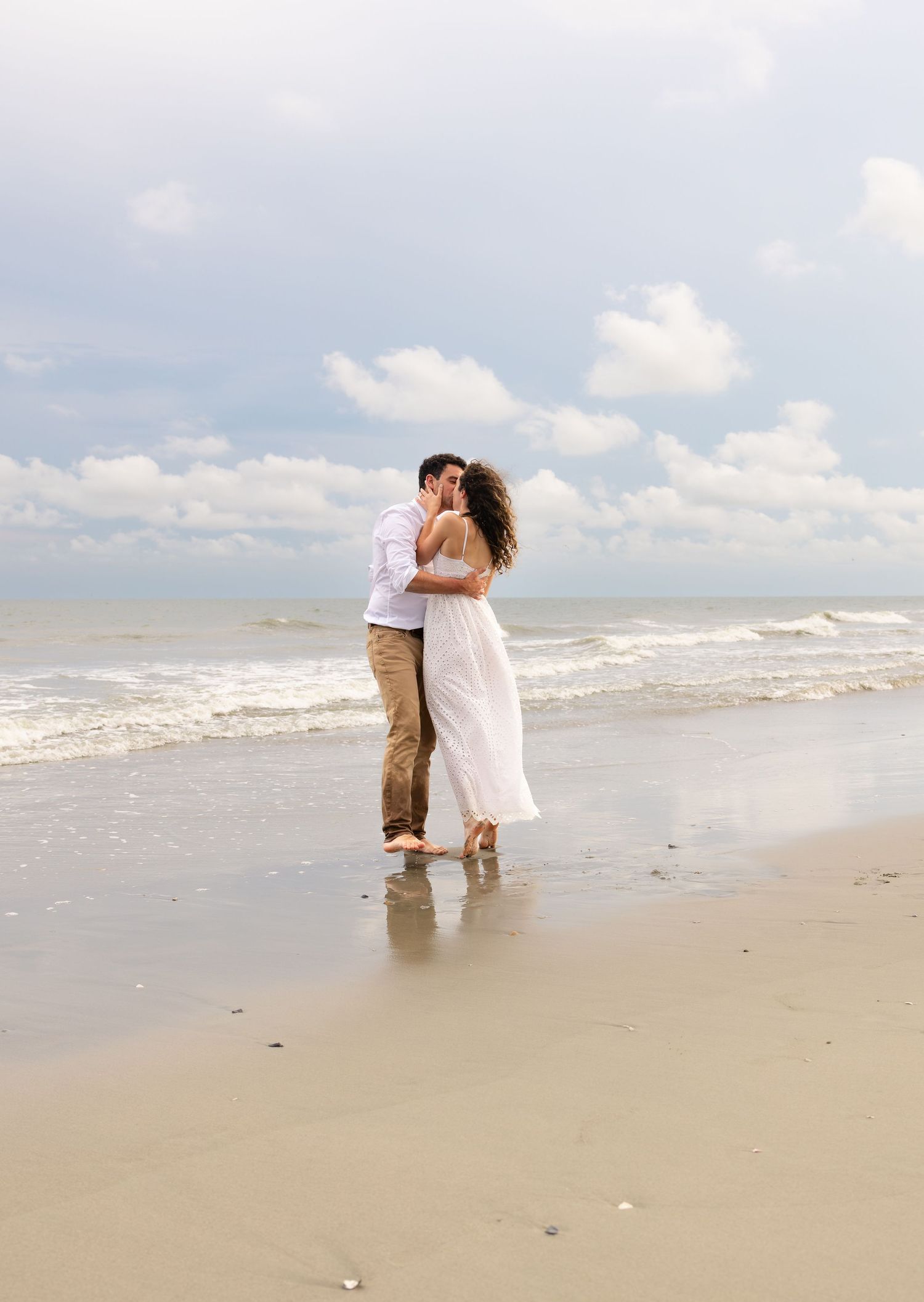 Romantic couple kissing on a sandy beach with ocean waves and cloudy sky in the background.