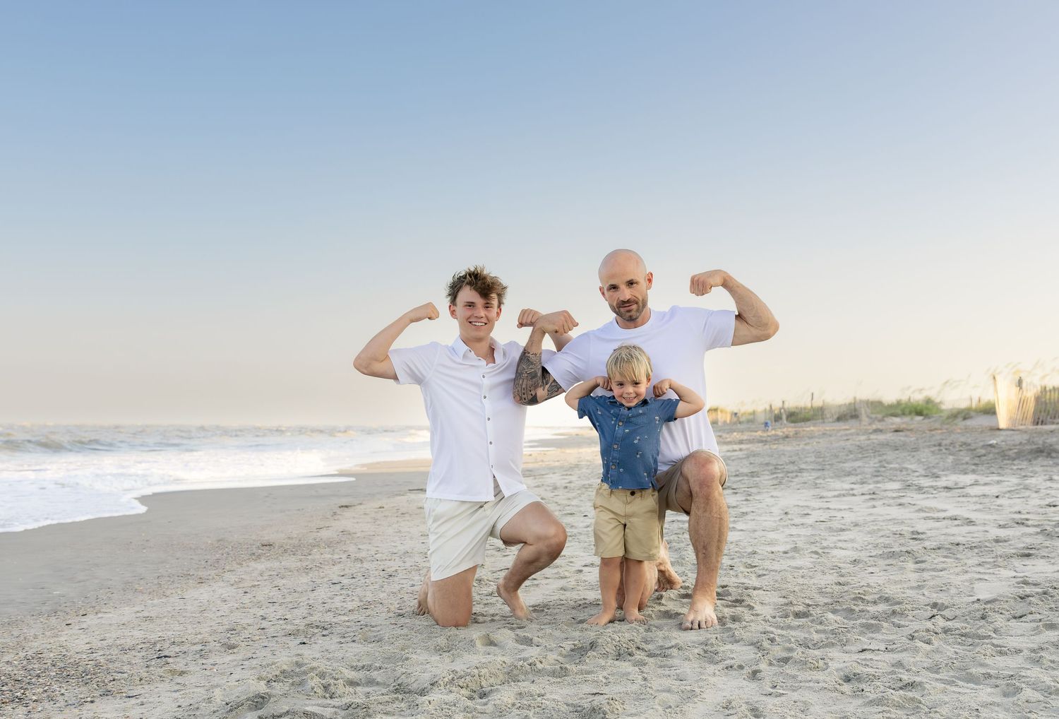Two boys and a man flexing muscles on a sandy beach with ocean waves in the background.