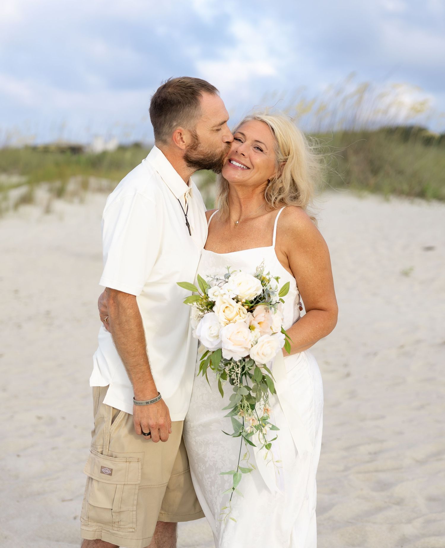 Couple in white attire sharing a romantic kiss on a sandy beach, woman holding a white floral bouquet.