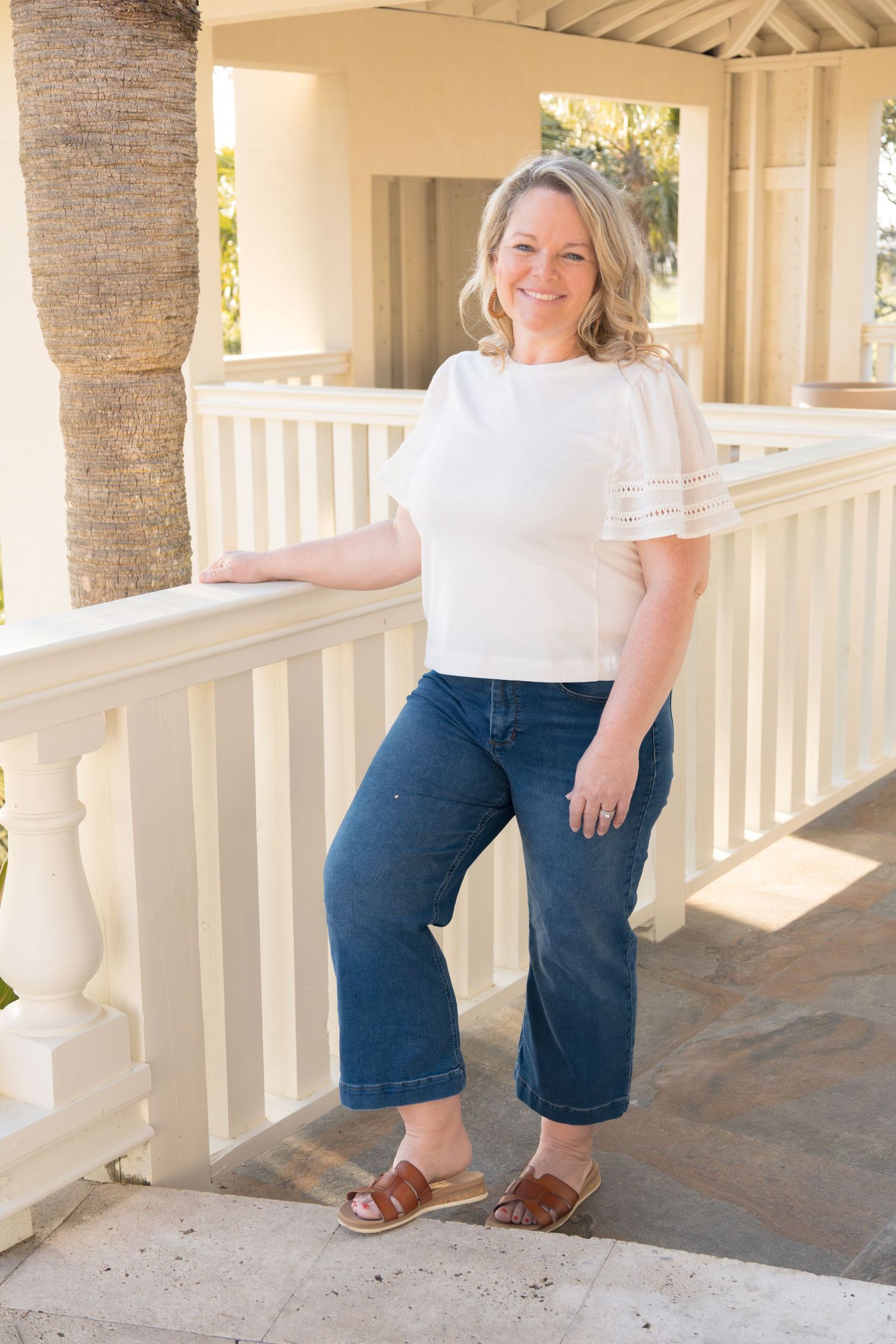 Smiling blonde woman in white top and wide-leg jeans poses on a sunny porch with white railings.