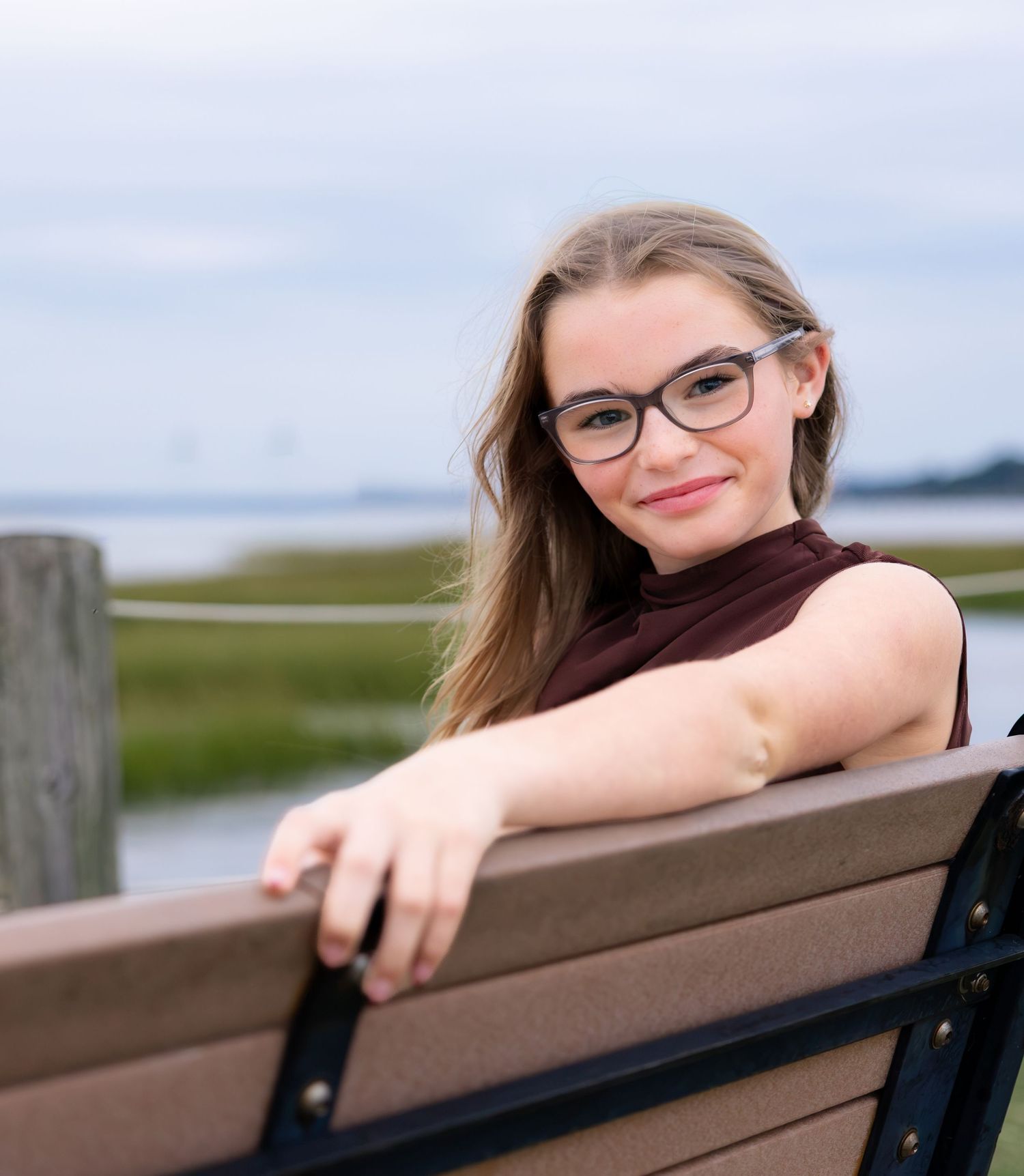 Young woman with glasses and long hair smiles on a waterfront bench in a sleeveless maroon top.
