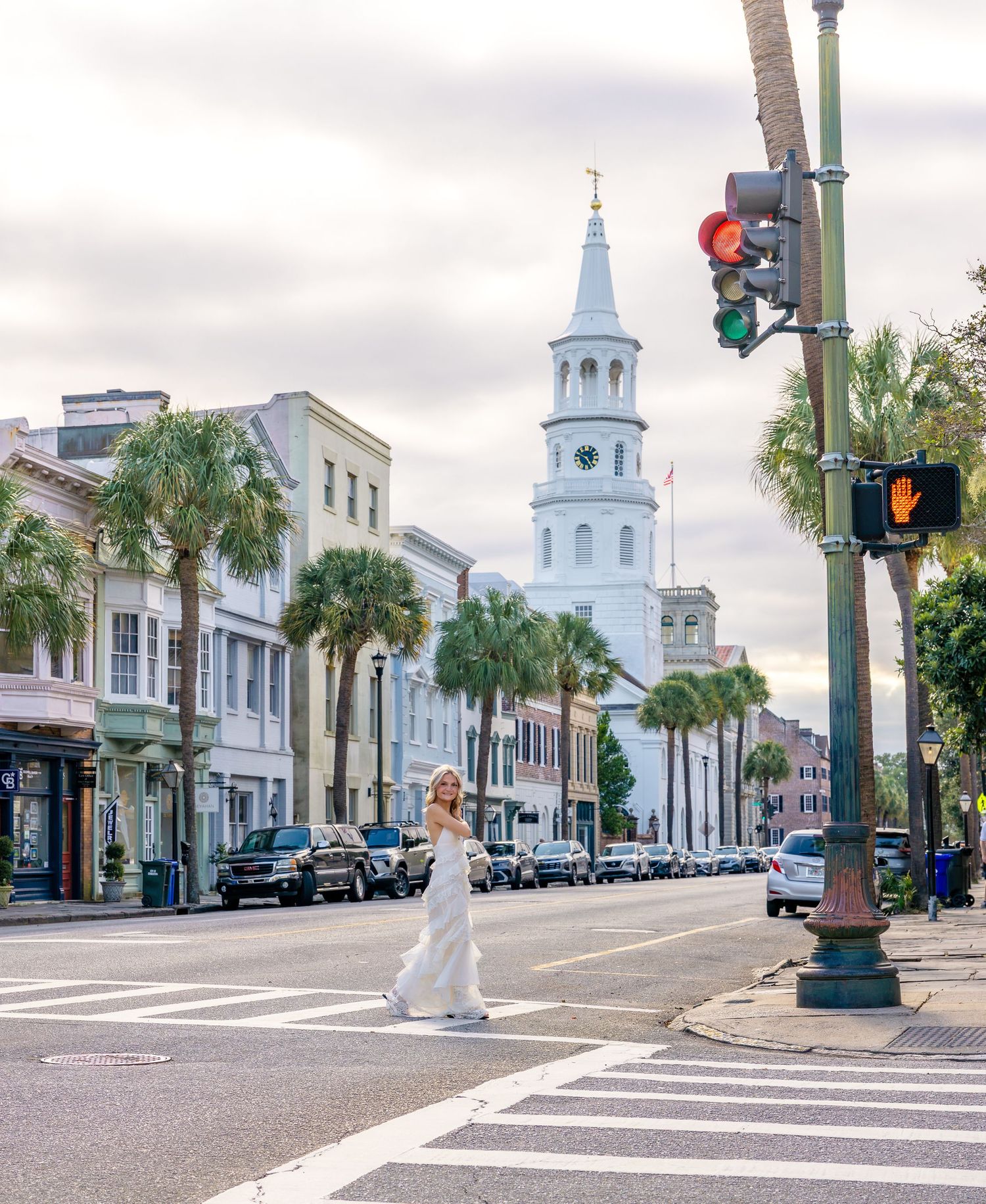 Woman in a white dress crosses a downtown street lined with palm trees, historic buildings and a white church steeple at sunset.