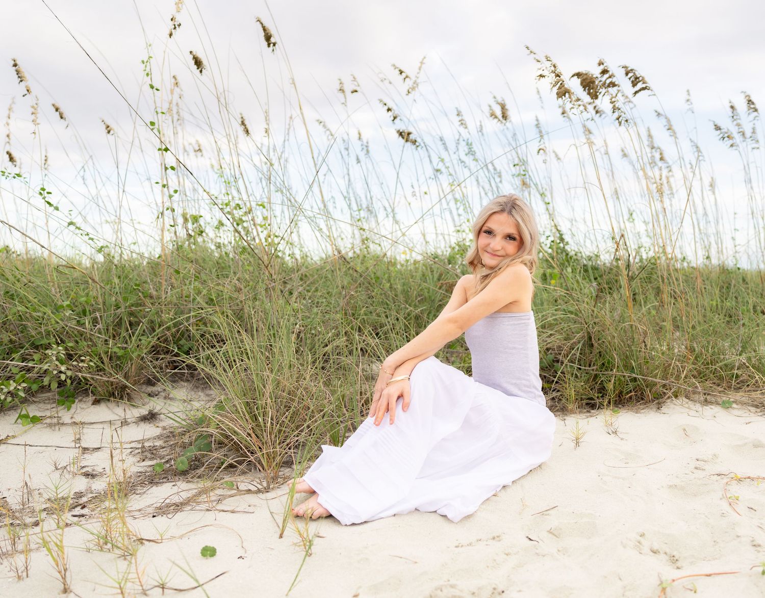 Blonde woman in a white dress sitting on a sandy beach dune surrounded by tall sea oats under a soft cloudy sky.