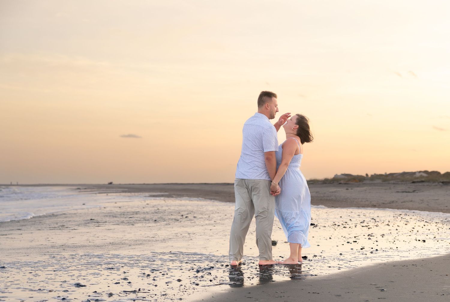 Couple in white outfits embracing on a wet sandy beach at sunset with soft orange sky and calm ocean waves in the background.