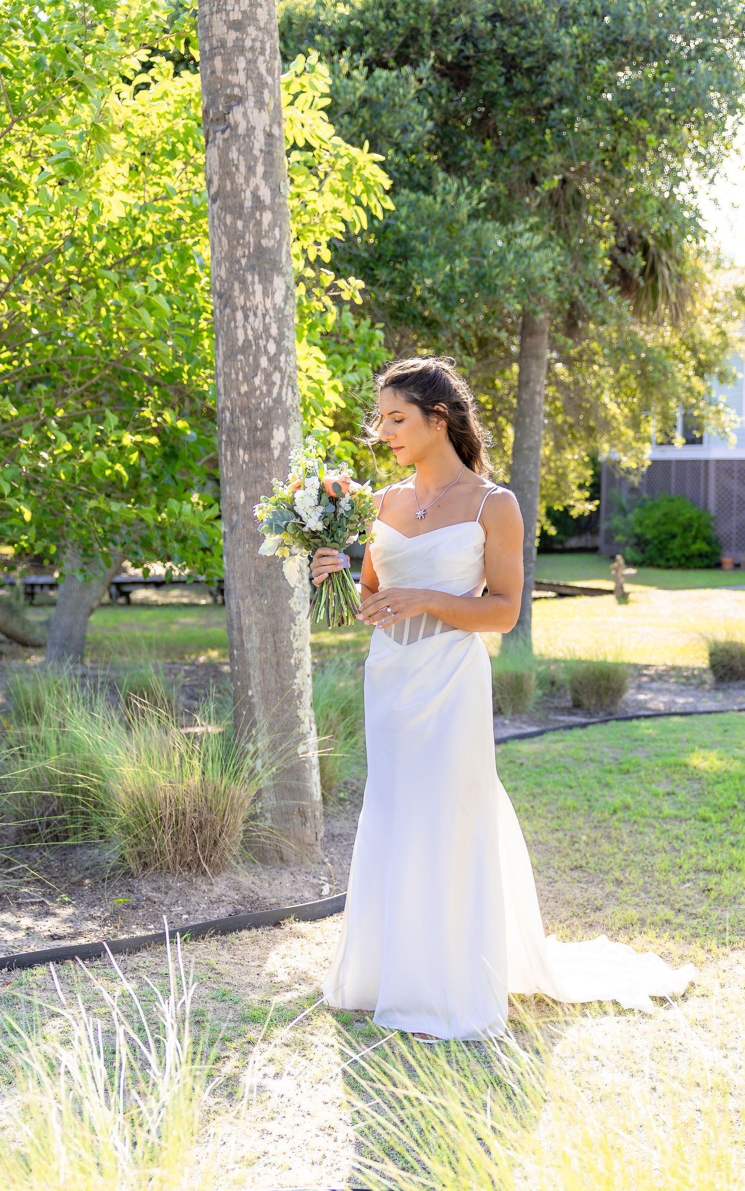 Bride in white spaghetti strap gown holding bouquet standing outdoors near trees on sunny wedding day.