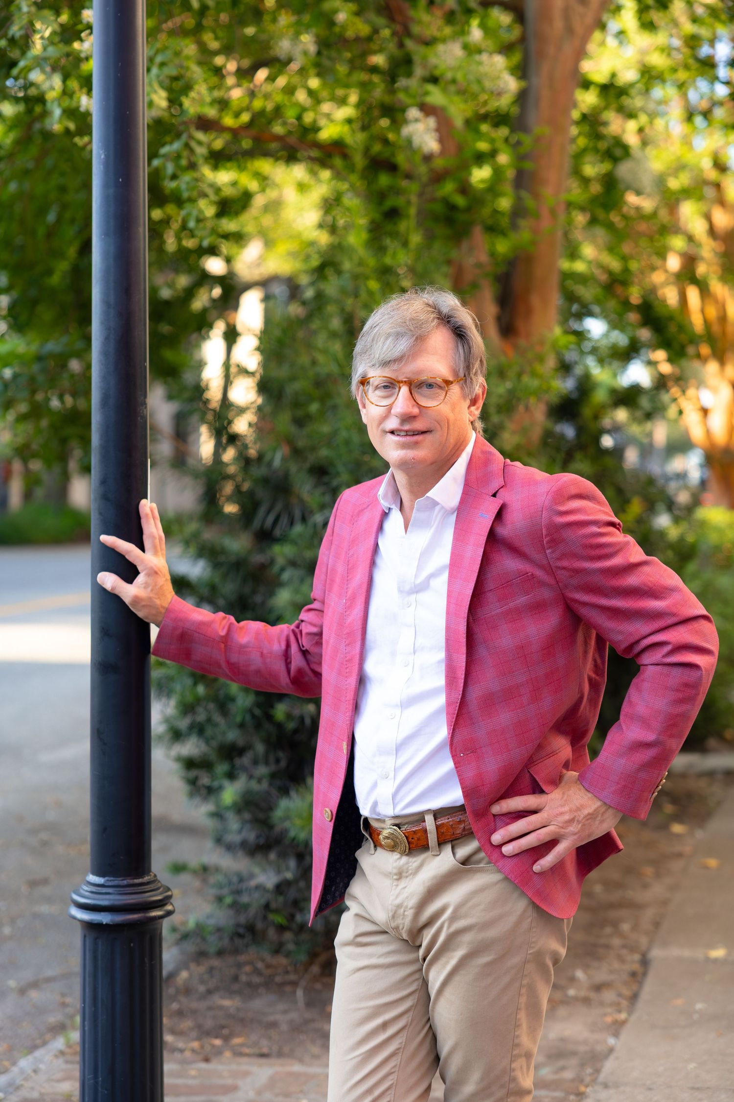 Middle-aged man in pink plaid blazer leaning on lamppost in sunny park setting.