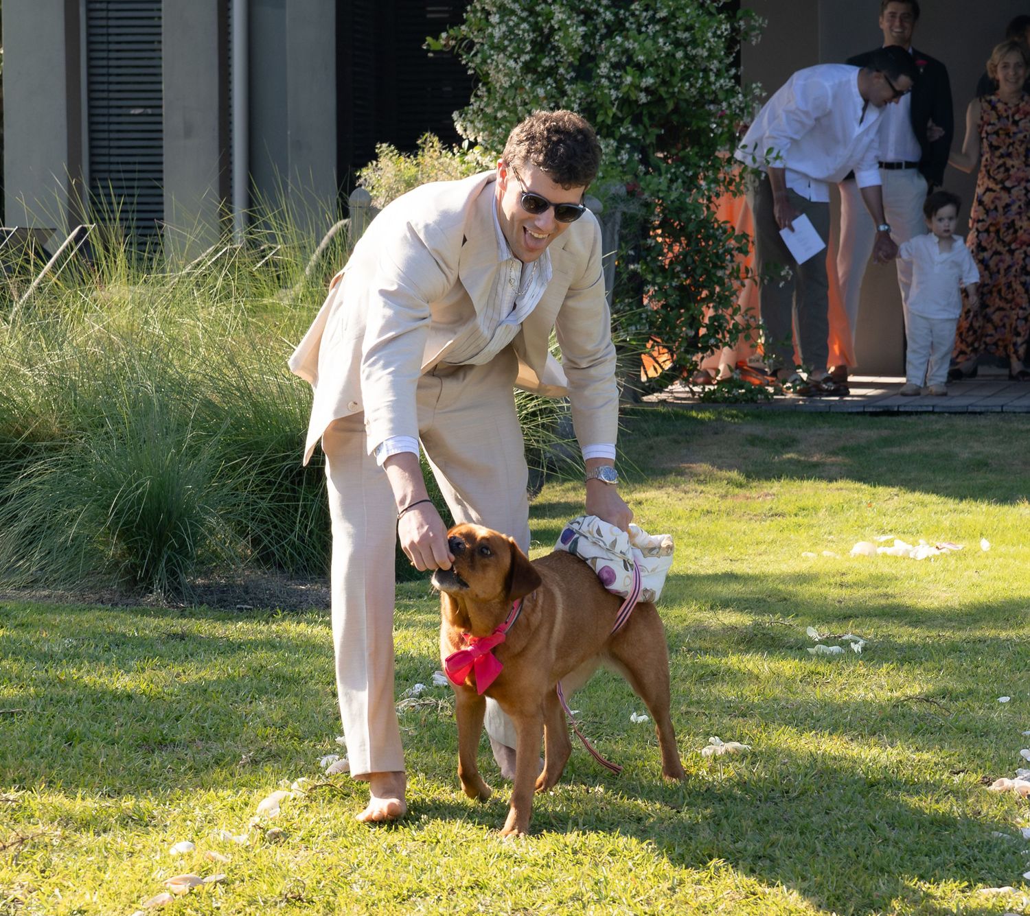 Man in beige suit petting a dog wearing a red bow tie on a green lawn at an outdoor wedding.