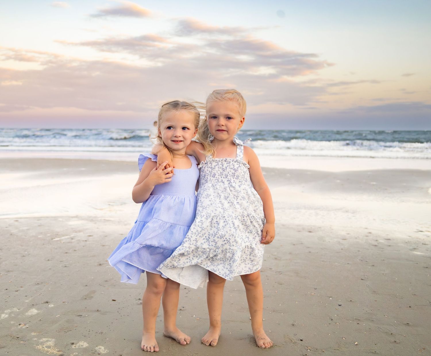 Two young girls in summer dresses pose together on a sandy beach at sunset with ocean waves behind them.