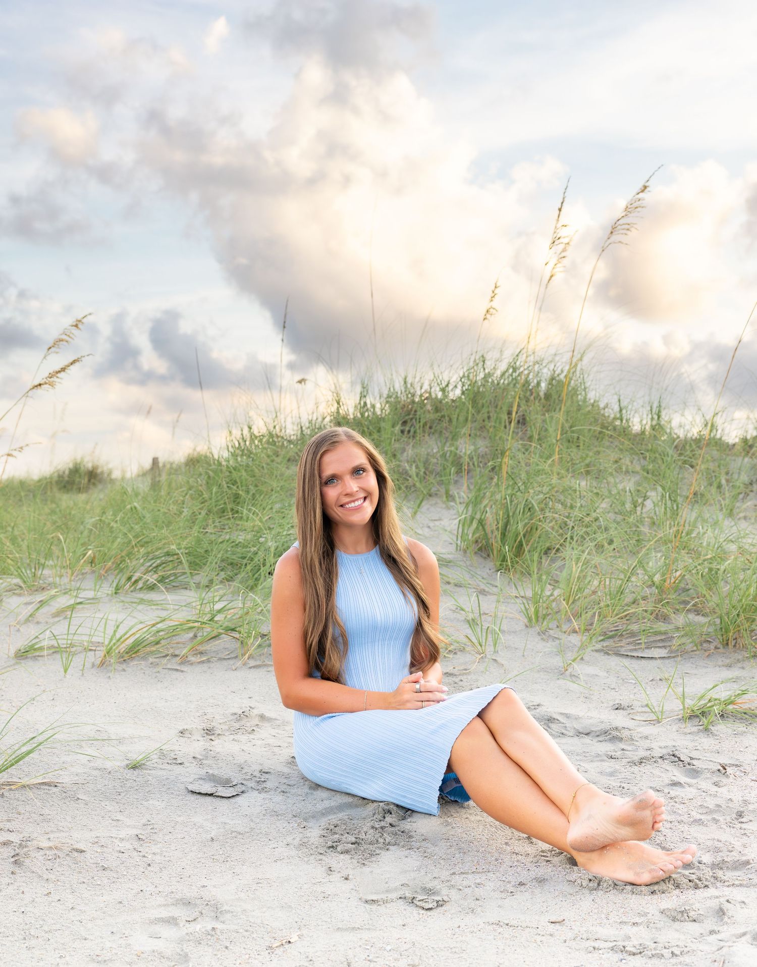 Young woman in light blue dress sitting barefoot on sandy beach dunes with sea oats and cloudy sky backdrop.