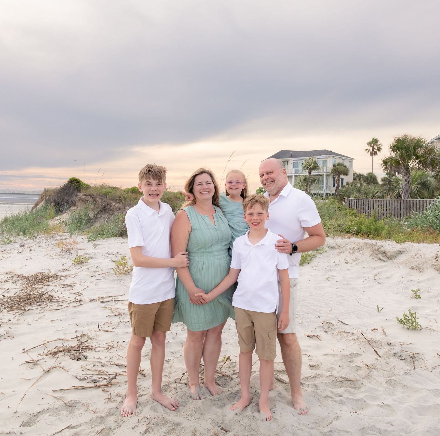 Family of five posing together on a sandy beach at sunset with a coastal house and palm trees in the background.