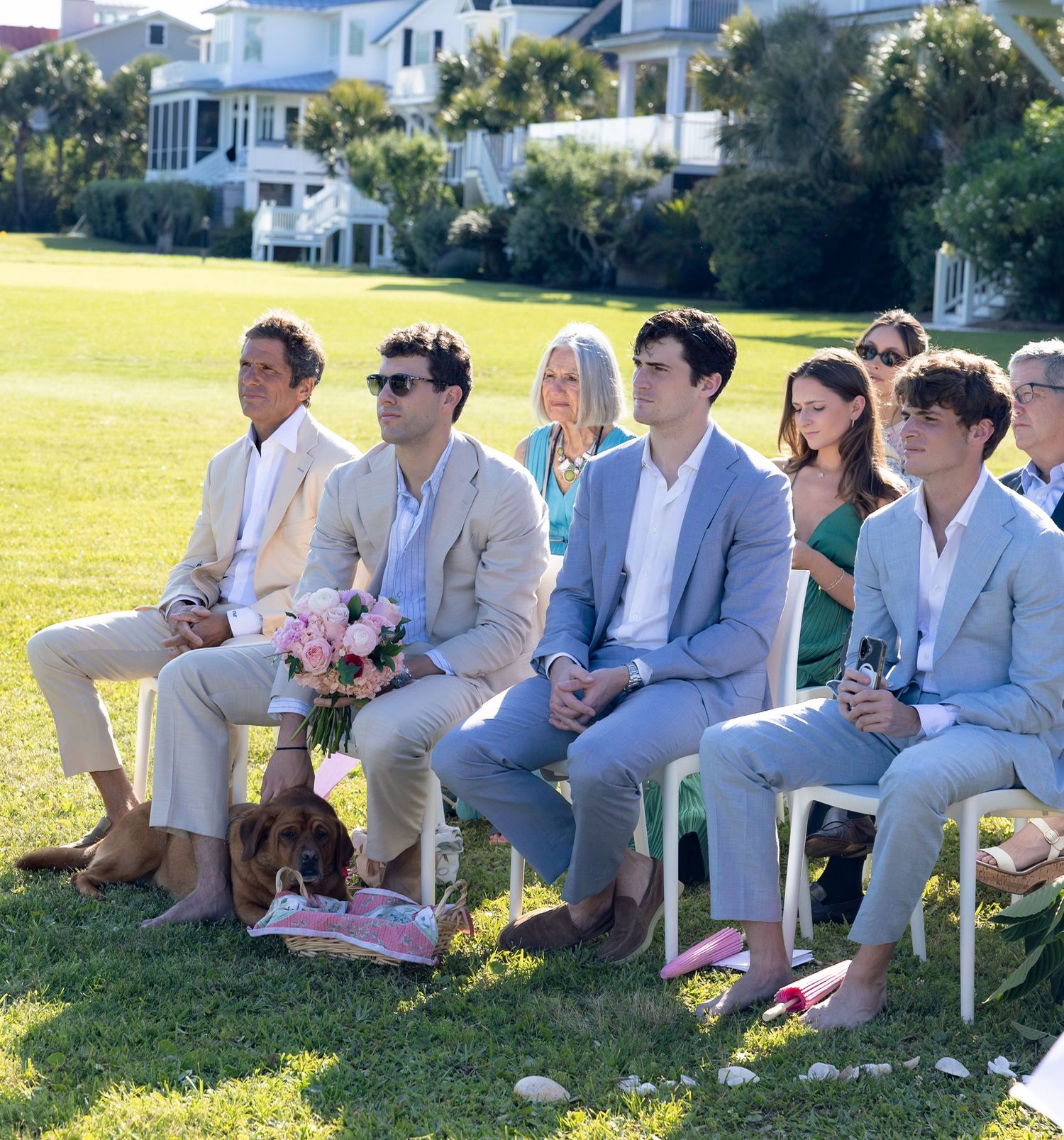 Wedding guests in light suits and dresses sit on white chairs on a sunny lawn, with a dog and a pink bouquet nearby, beside a coastal home.