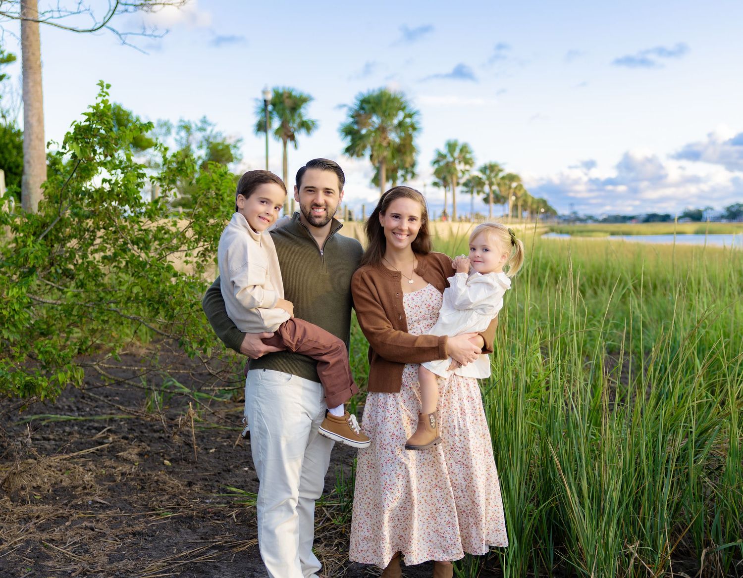 Smiling family of four holding two young children while posing outdoors near tall marsh grass and palm trees at sunset.