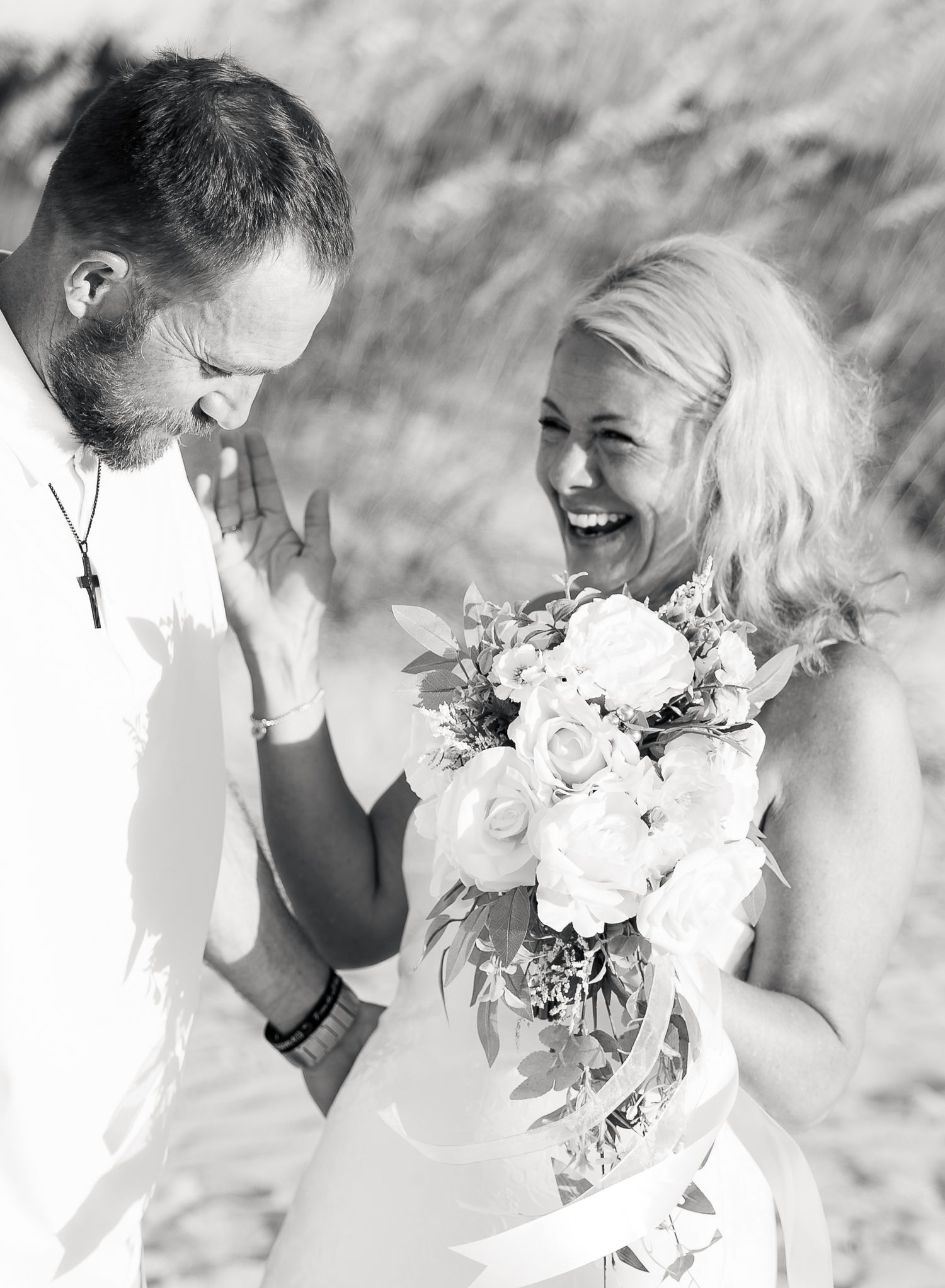 Black and white photo of a laughing bride holding a white rose bouquet with her groom on a beach wedding day.