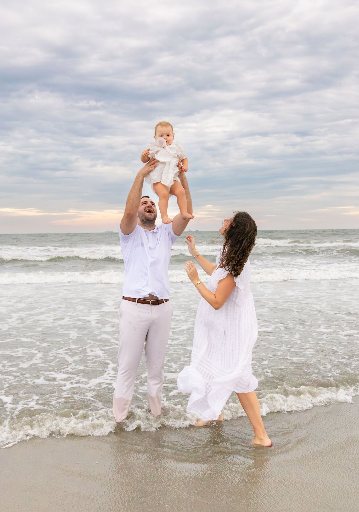 Family in white outfits playing on beach, father lifting baby while mother reaches up, ocean waves in background.
