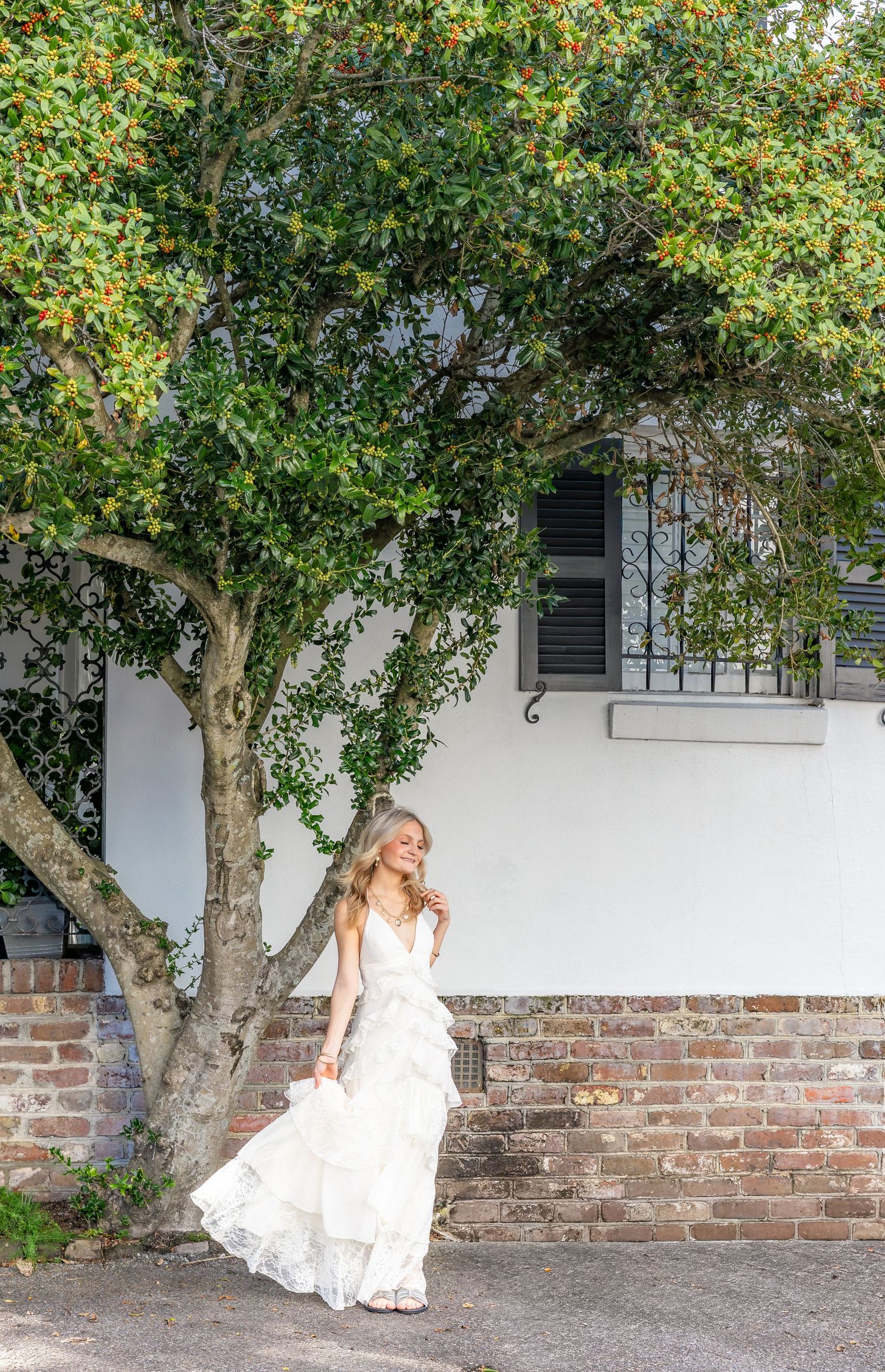Woman in a white tiered ruffle dress standing beneath a leafy tree beside a white stucco house with a brick foundation and black shutters.