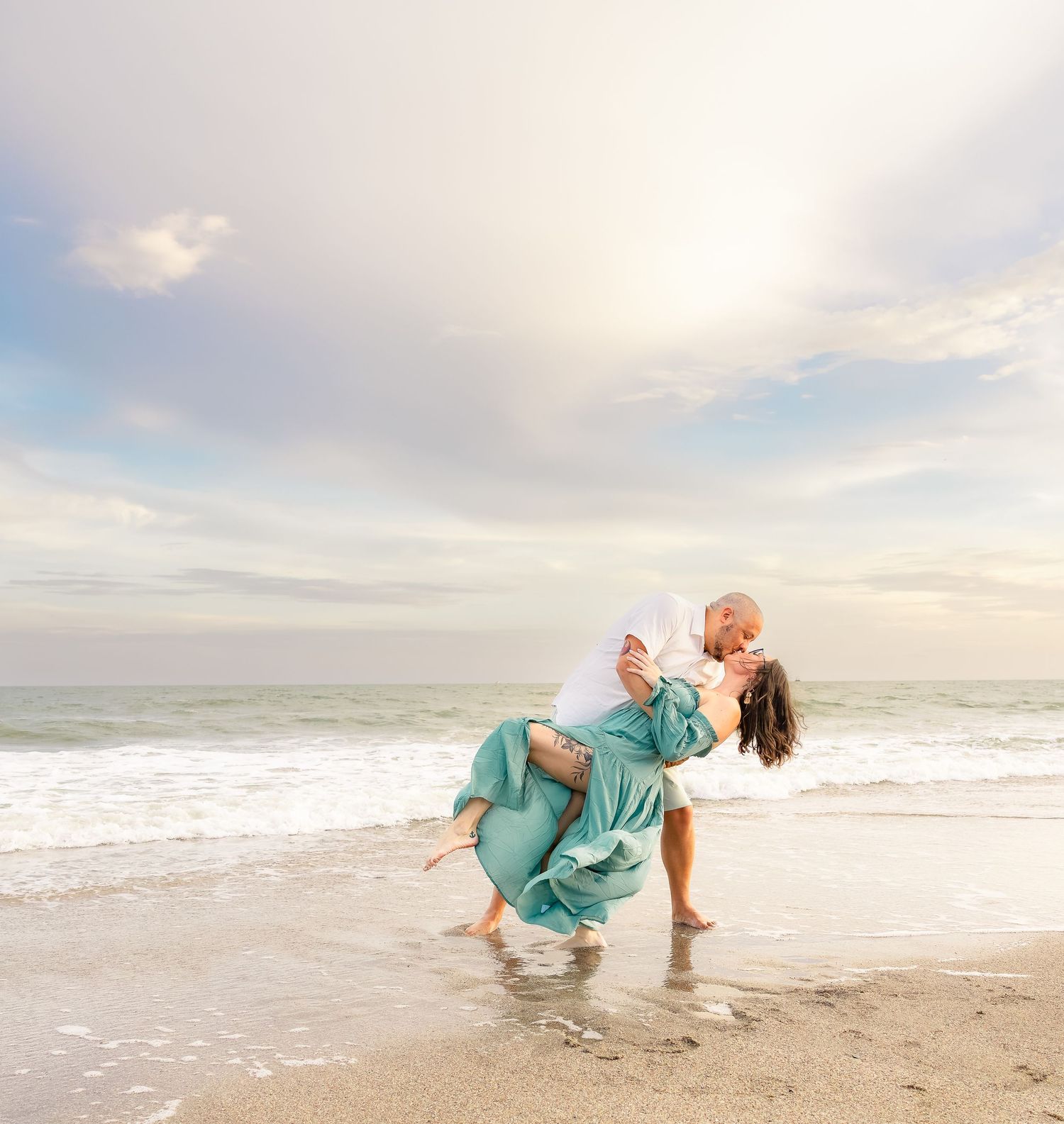 Couple in a romantic dip kiss on a sandy beach at sunset, her flowing teal dress and the ocean waves behind them.