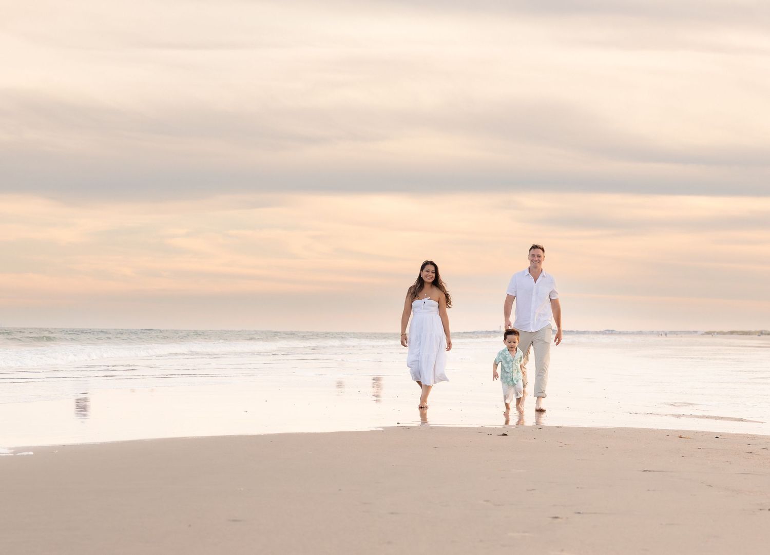 Family of three walking on a sandy beach at sunset, parents and young child holding hands in white outfits.