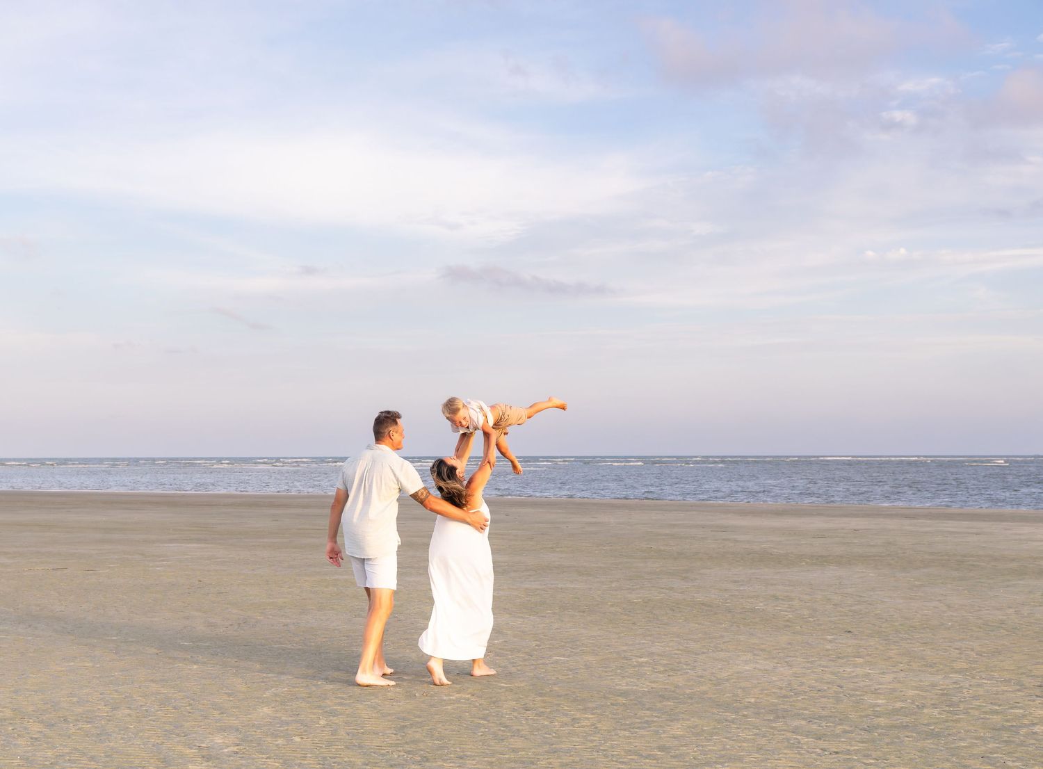 Family of three enjoying a beach walk at sunset, parents lifting child in white outfits on sandy shore.