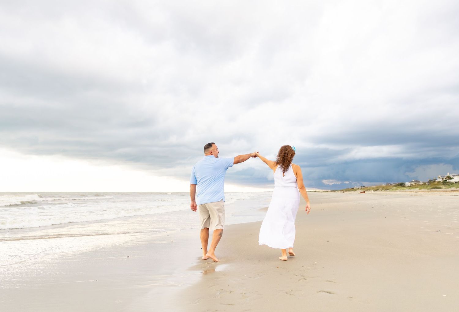 Couple holding hands dancing on a sandy beach with dramatic cloudy skies and ocean waves in the background.