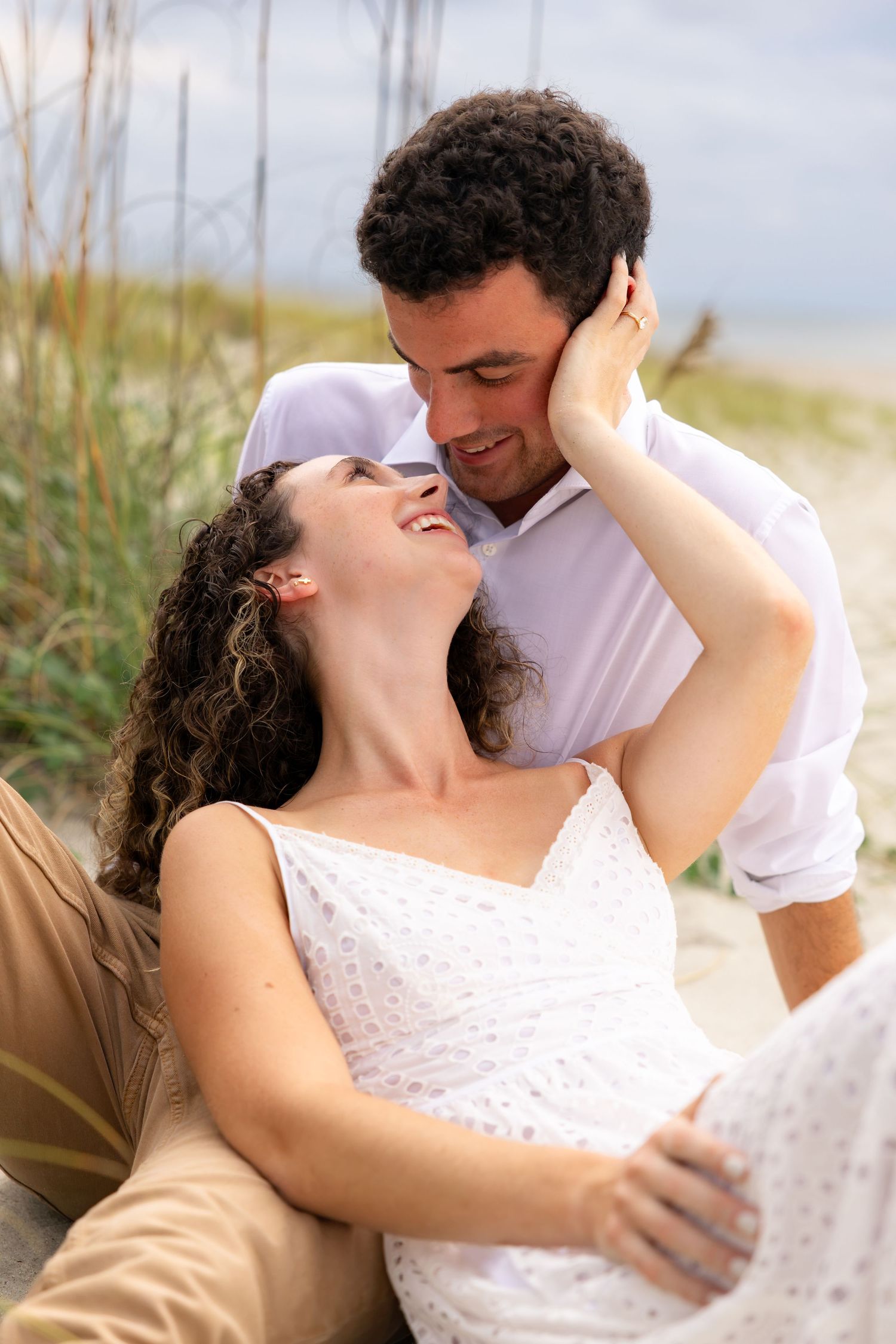 Romantic couple sharing a tender moment on a sandy beach, woman in white dress smiling up at man in white shirt.