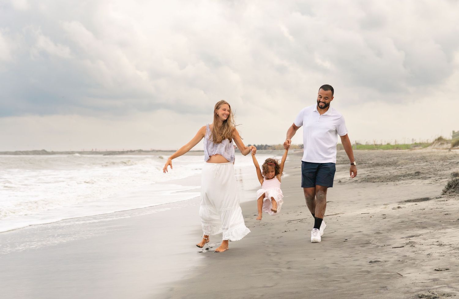Family of three walking on beach, parents swinging toddler between them, ocean waves in background.