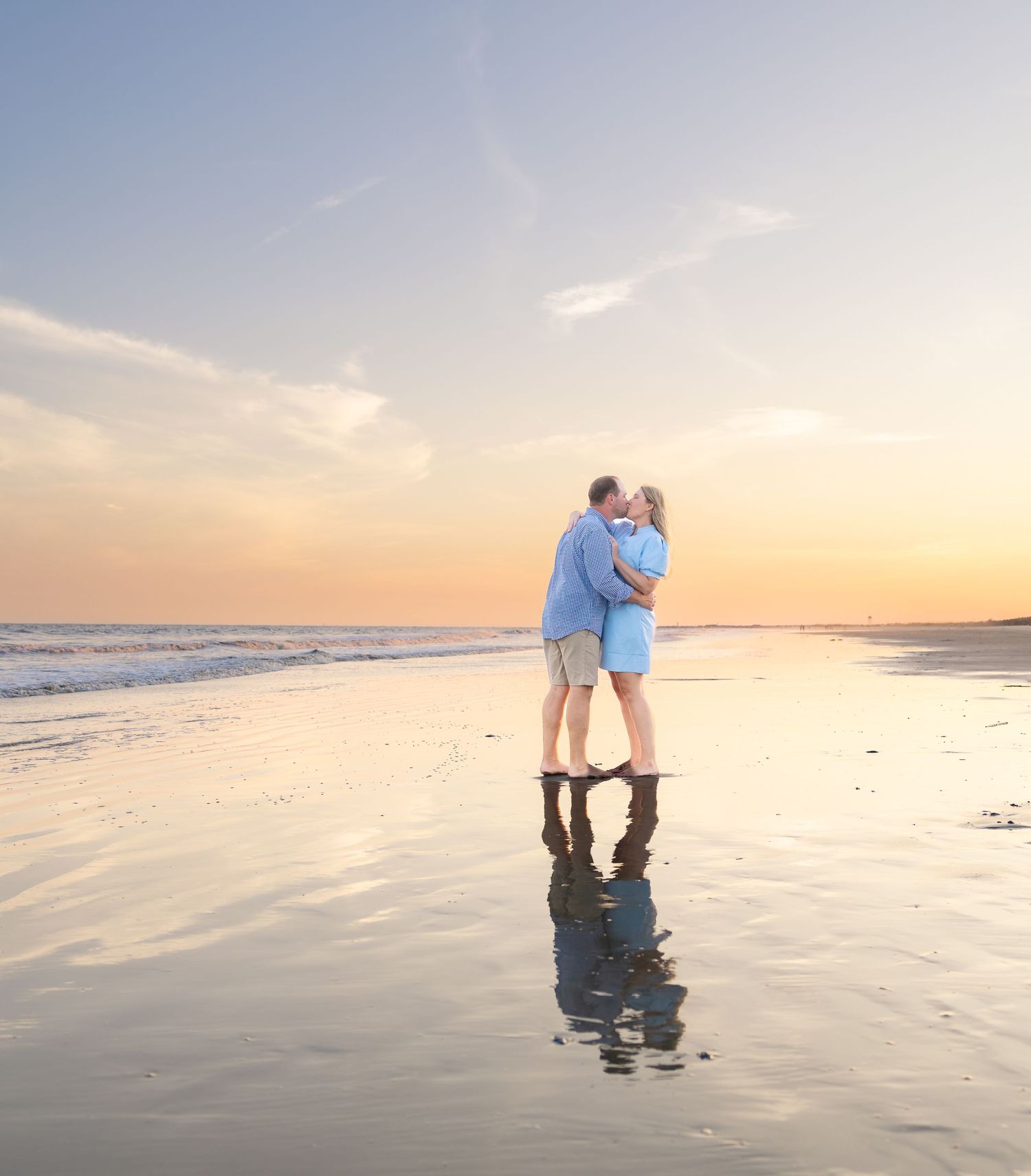 Couple embracing and kissing on a reflective beach at sunset with golden skies and ocean waves.